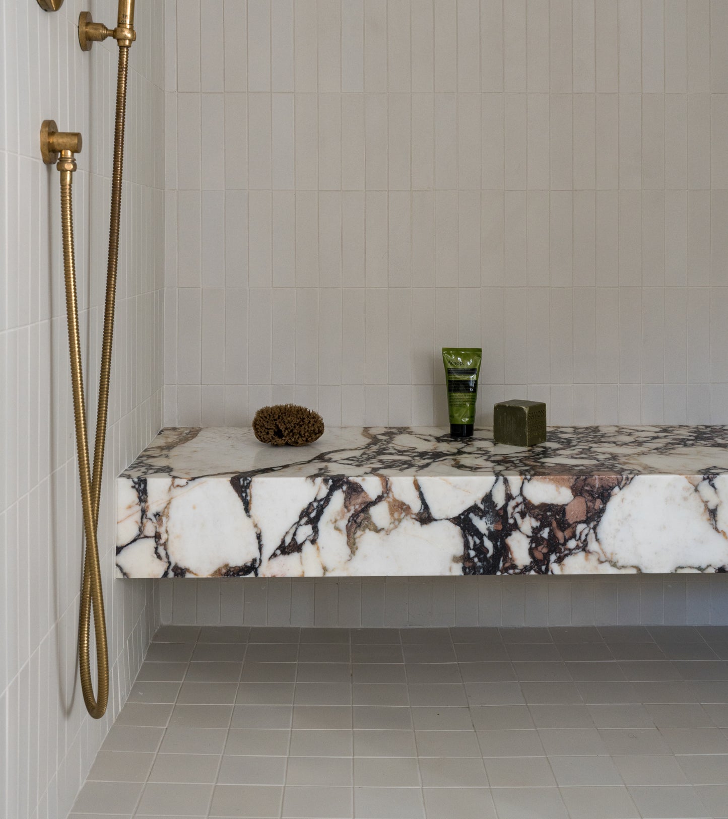 A modern shower features gold fixtures, a marble bench with accessories, and Zia Tile's Linen 4x4 ceramic tiles on the walls, paired with a matte ceramic tile floor.