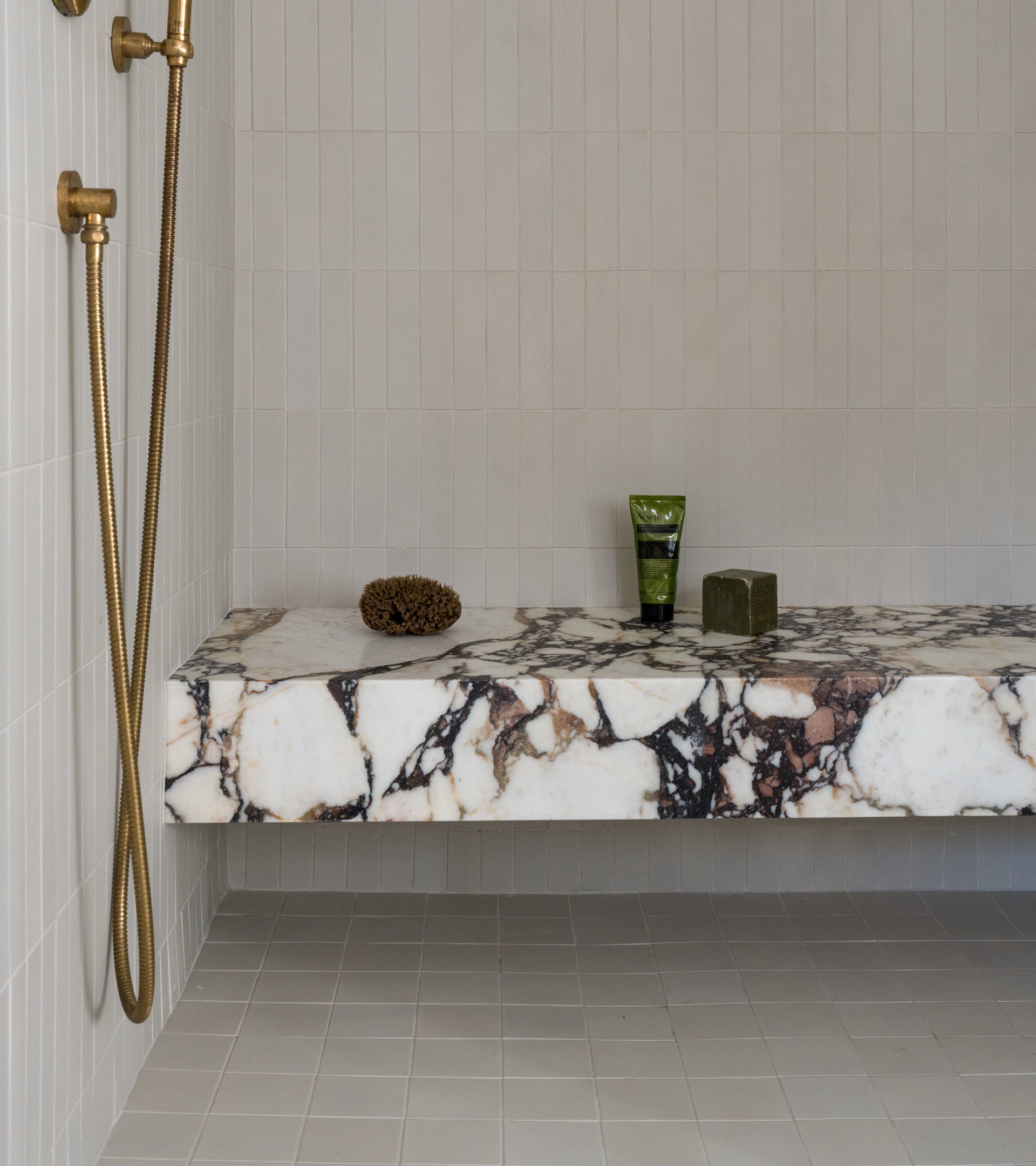 A modern shower features gold fixtures, a marble bench with accessories, and Zia Tile's Linen 4x4 ceramic tiles on the walls, paired with a matte ceramic tile floor.