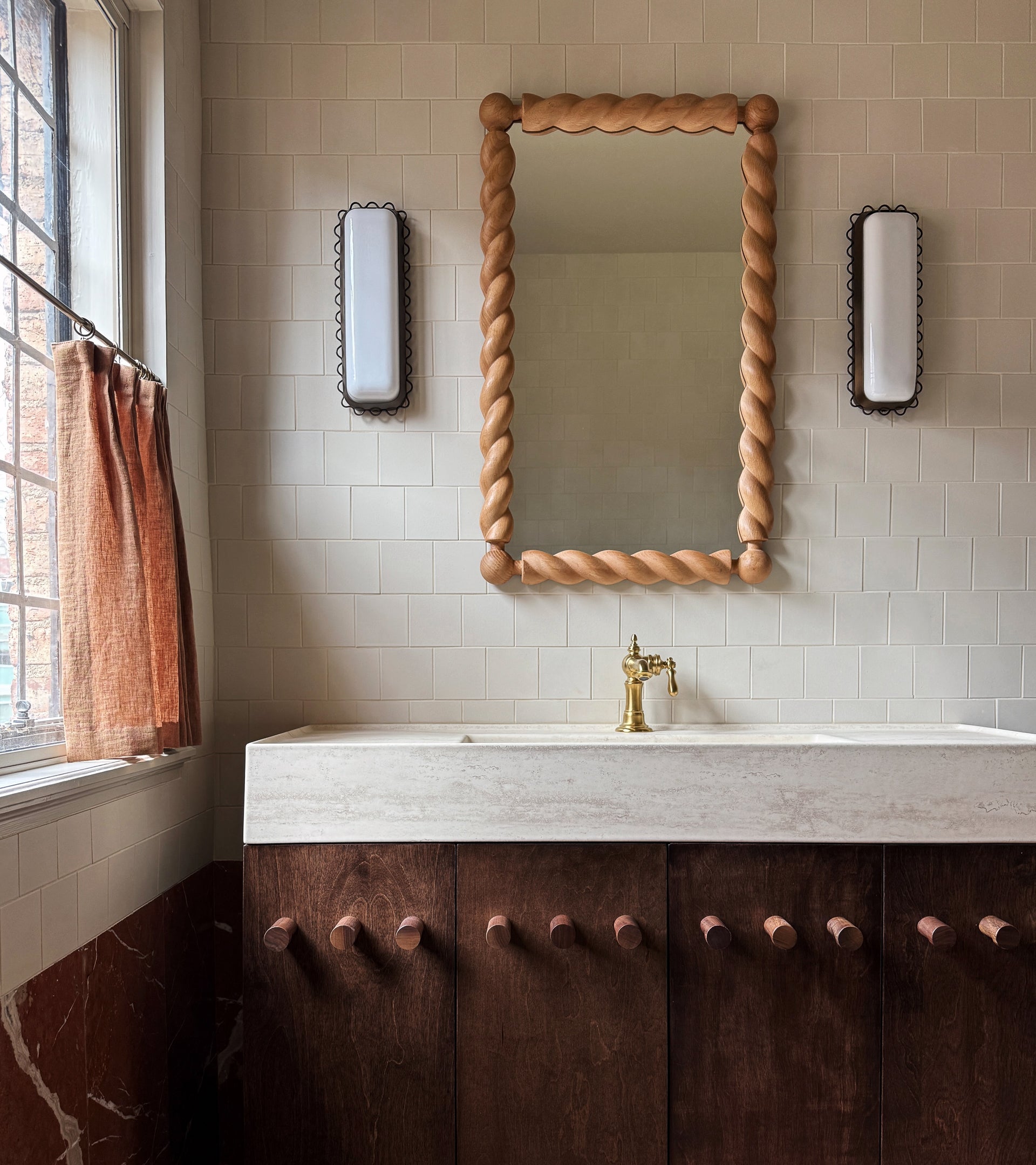 A bathroom features a twisted wood-framed mirror above a marble countertop with brass faucet, dark wooden vanity, and Linen 4x4 ceramic tiles from Zia Tile beneath a window dressed in rust-colored curtains, with wall lights flanking the mirror.