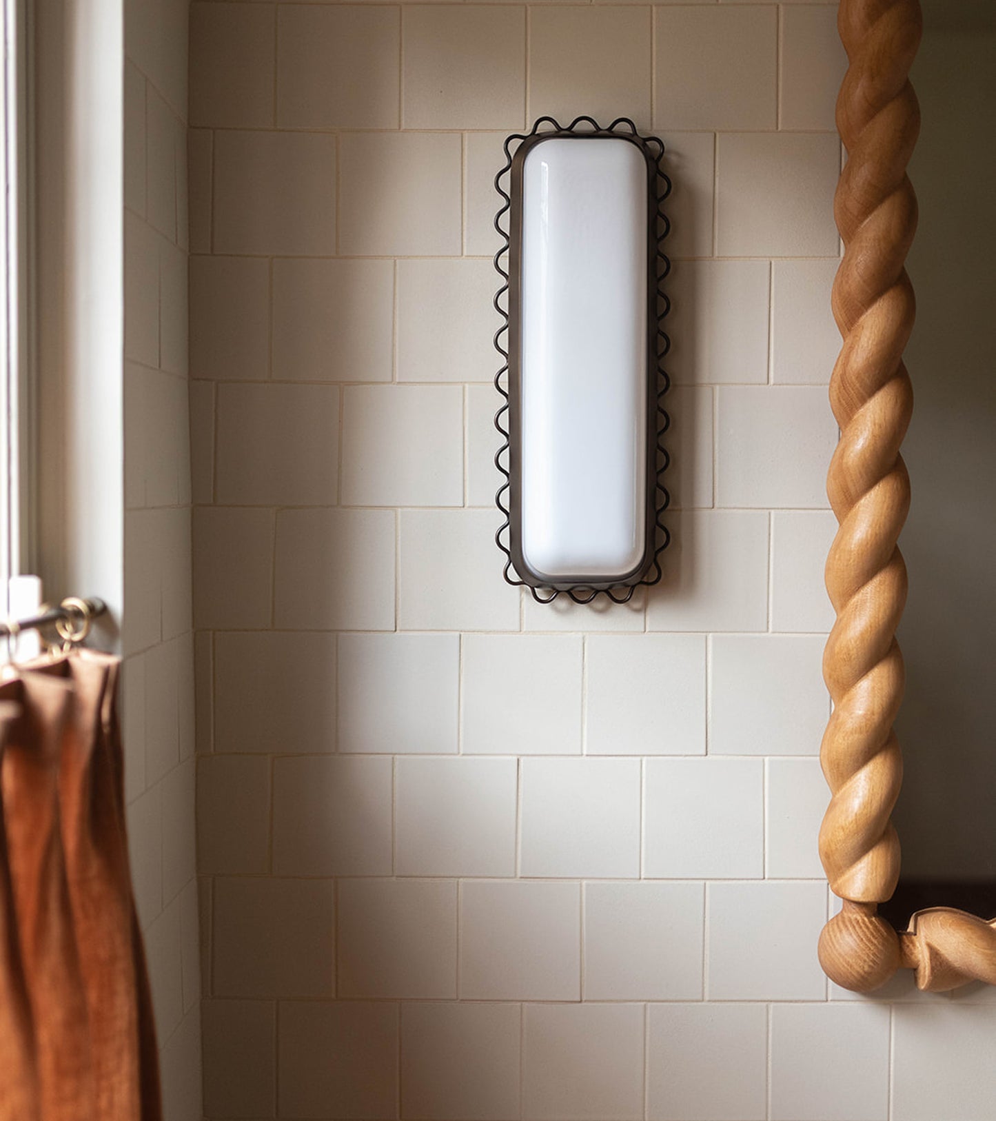 A rectangular white wall sconce with a black scalloped frame is mounted on a bathroom wall clad in Zia Tile’s Linen 4x4 matte ceramic tile. A mirror with a thick twisted wooden frame and a brown curtain are also visible.