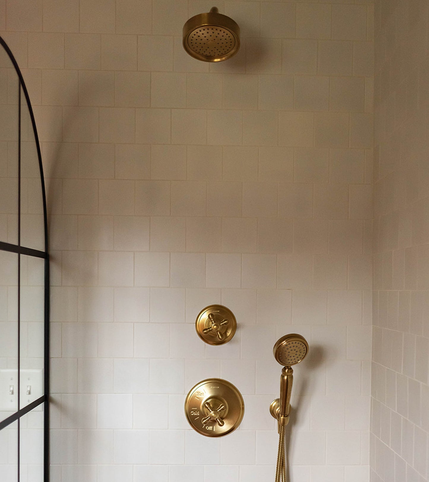 A modern shower features Zia Tile’s Linen 4x4 white ceramic tiles, gold fixtures (showerhead, handheld sprayer, control knobs), and a black-framed glass shower door partially visible on the left.