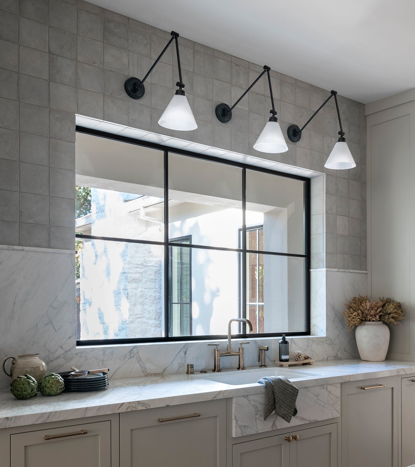 Modern kitchen featuring a Monument 6x6 + Honed marble countertop and backsplash, large black-framed window, beige cabinets, brass fixtures, wall lights, plus decorative plates and dried flowers. Large format stone tiles enhance the bright space.