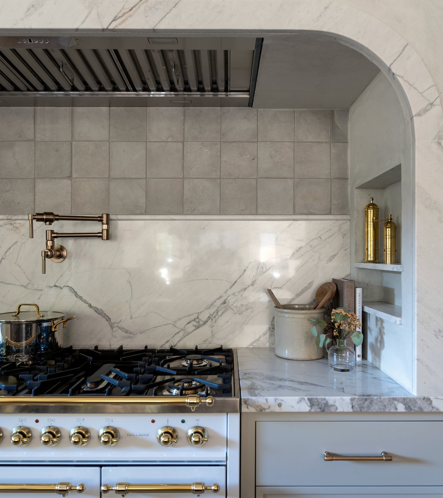 A modern kitchen showcases a Monument 6x6 + Honed marble countertop and backsplash, large format stone accents, a brass-knob gas stove, pot filler faucet, saucepan, decorative jars, flower vase, and built-in shelving.