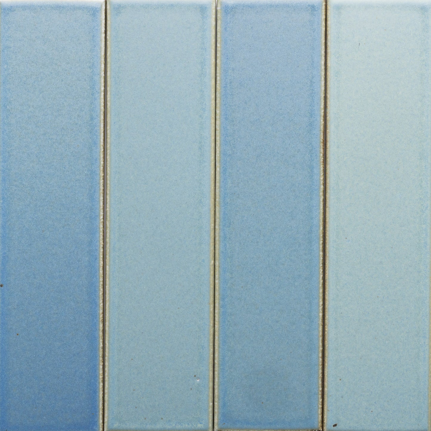 Four vertical rectangular Morning Light + Flat tiles by Zia Tile, in varying blue shades, are arranged side by side with thin, slightly raised grout lines. The tiles have a smooth, matte surface.