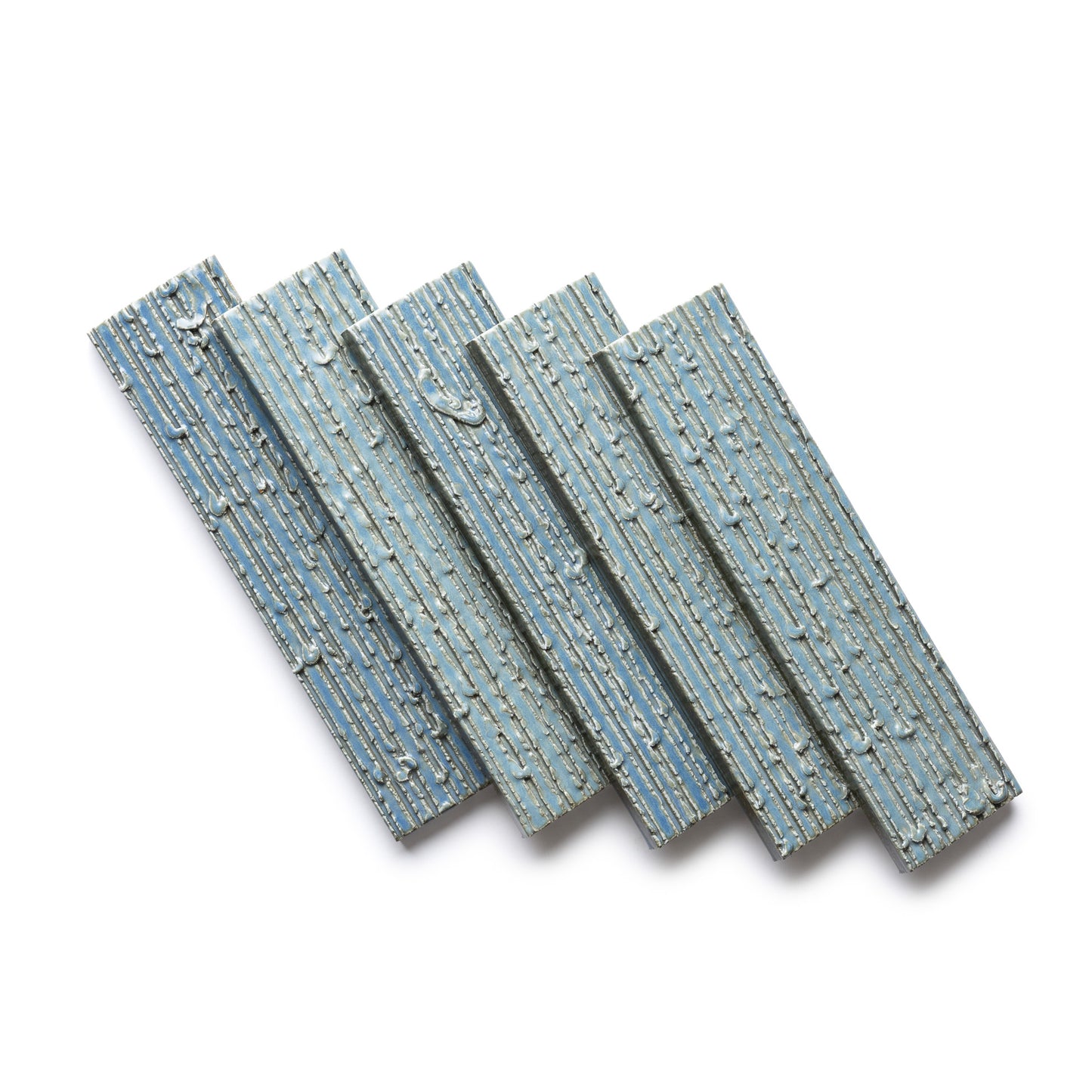 Four “Morning Light + Furrowed” tiles by Zia Tile, in textured blue-gray ceramic, are arranged diagonally on a white background. Each features an embossed linear pattern reminiscent of bark or wood grain.