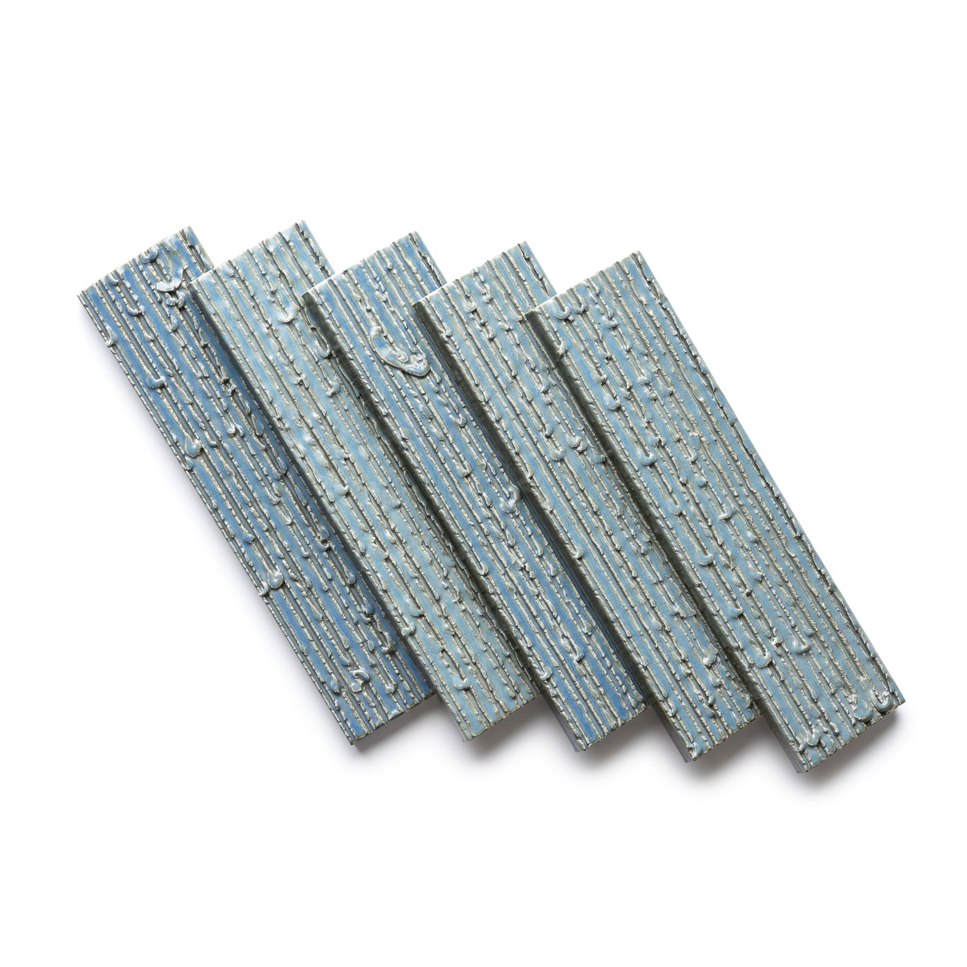 Four “Morning Light + Furrowed” tiles by Zia Tile, in textured blue-gray ceramic, are arranged diagonally on a white background. Each features an embossed linear pattern reminiscent of bark or wood grain.