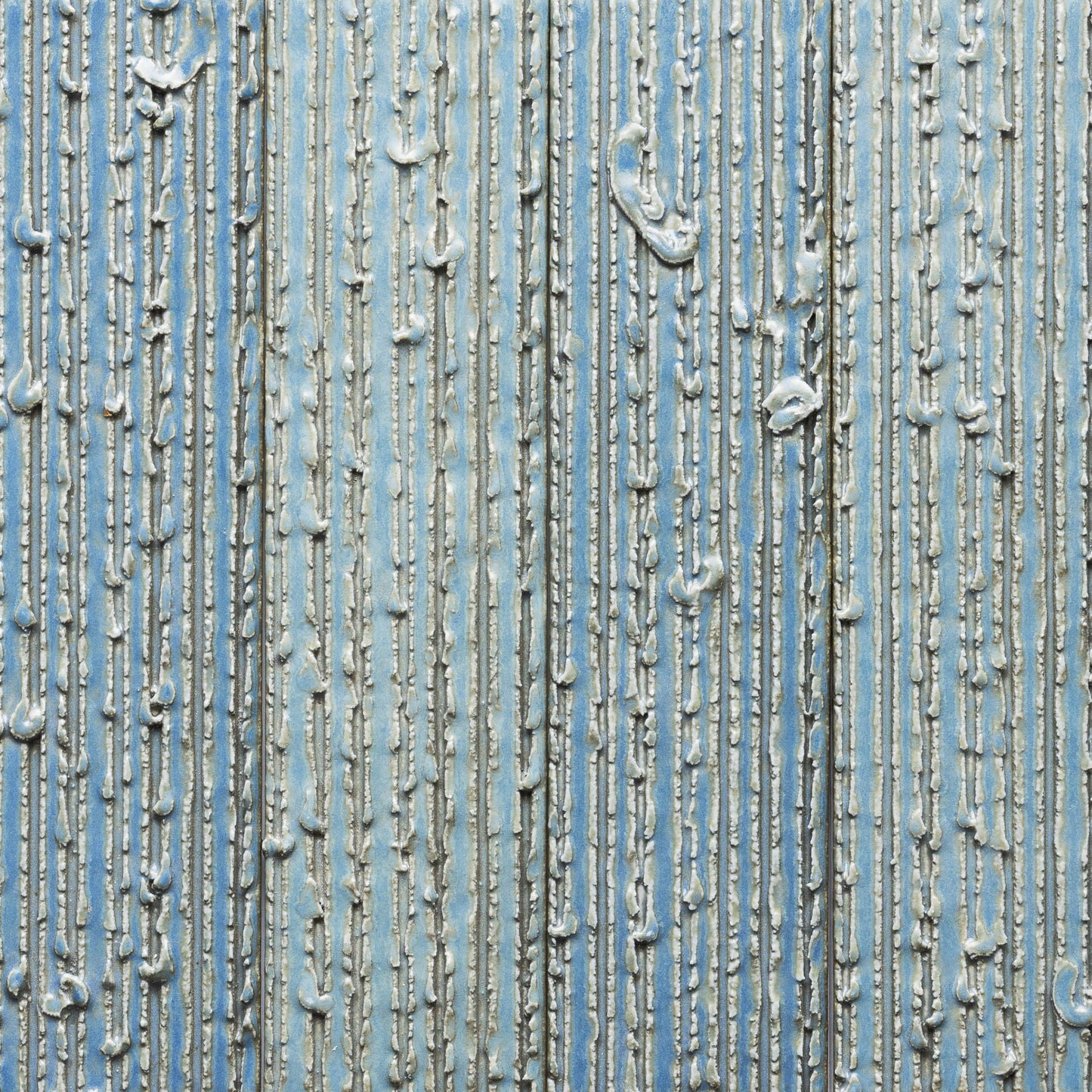Close-up of Zia Tile's Morning Light + Furrowed tile, showing vertical silver ridges and raised, uneven lines on a blue background for a tactile, abstract effect.