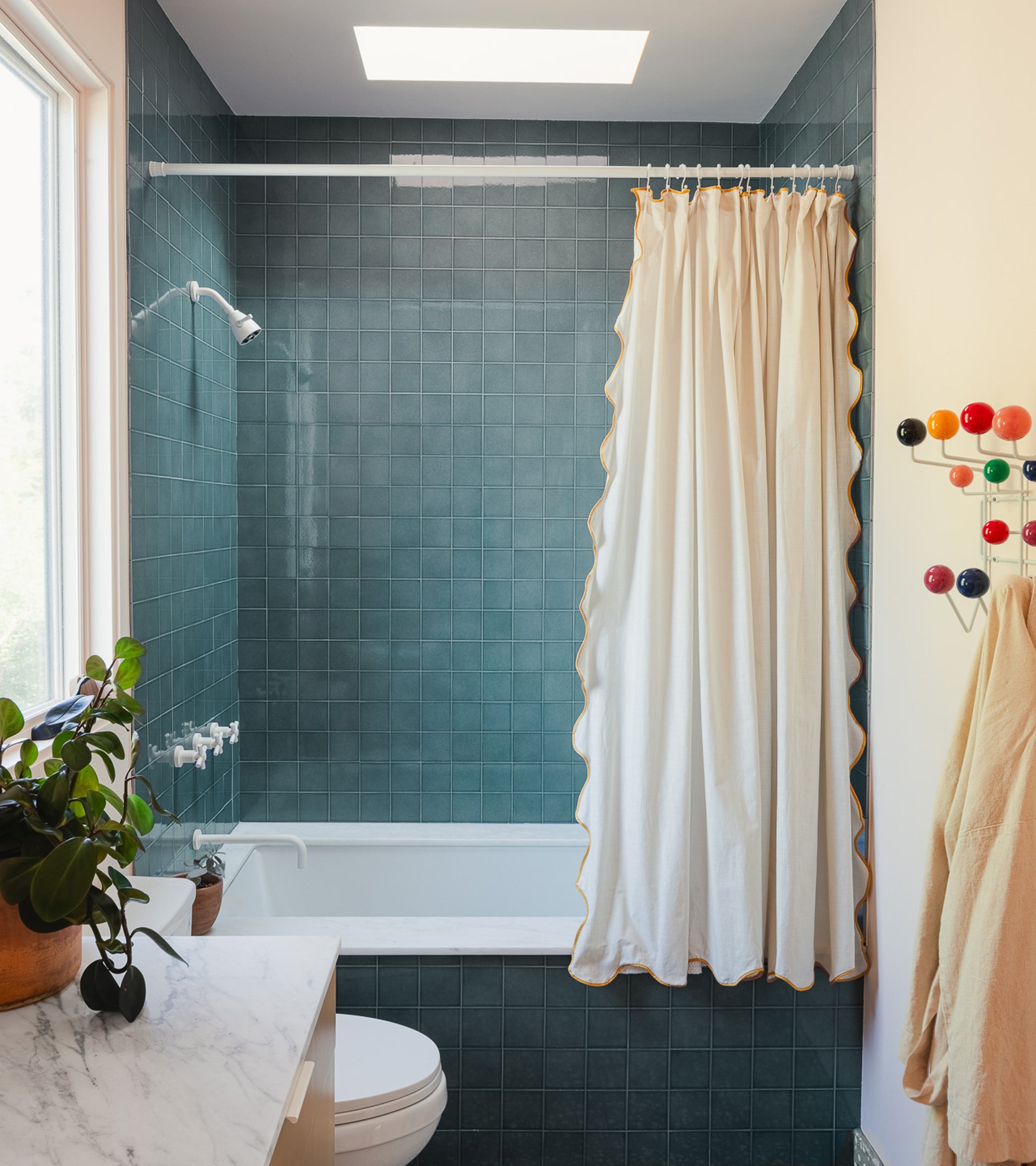 A small bathroom featuring Zia Tiles Nagano 4x4 glossy teal ceramic tile, a white bathtub, white-yellow trimmed shower curtain, white toilet, a towel on a hook, potted plant on marble counter, and colorful wall hooks.