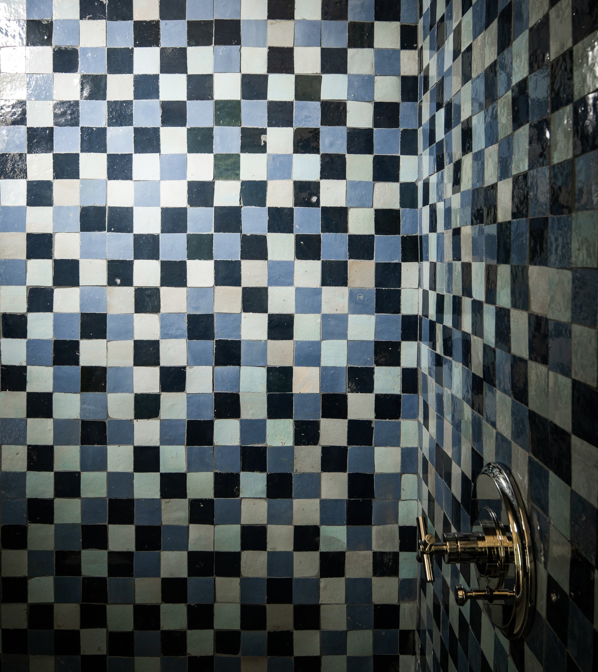 A shower wall features Zia Tile’s Night Blue + Skylight + Portuguese Blue Checkerboard tiles in a glossy 2x2" pattern, accented by a silver shower handle on the lower right.