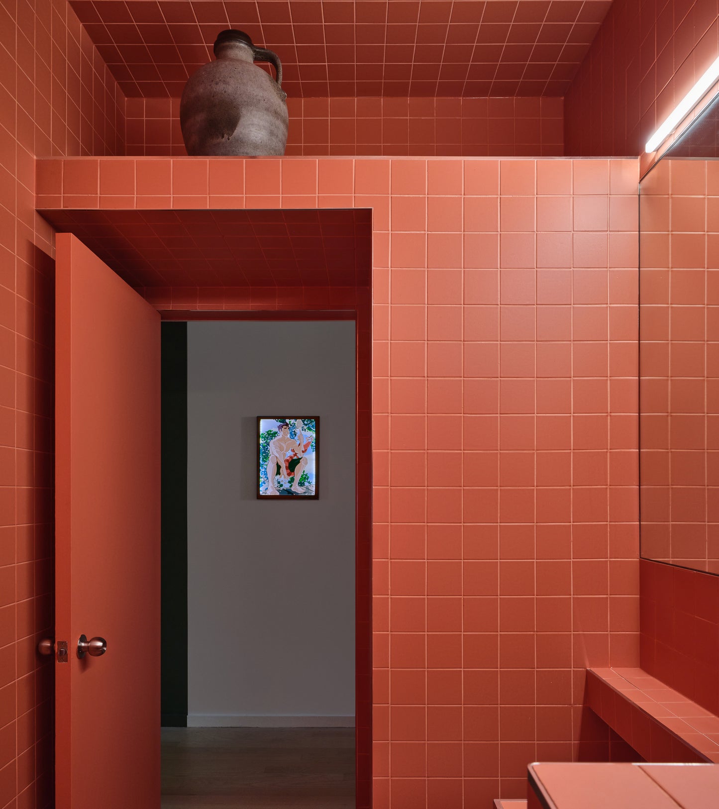 A modern bathroom features Zia Tile’s Pink Dahlia 4x4 matte ceramic tiles in red-orange, a matching open door, and a large ceramic jug on a shelf. A lit art piece is visible on a white wall through the doorway.