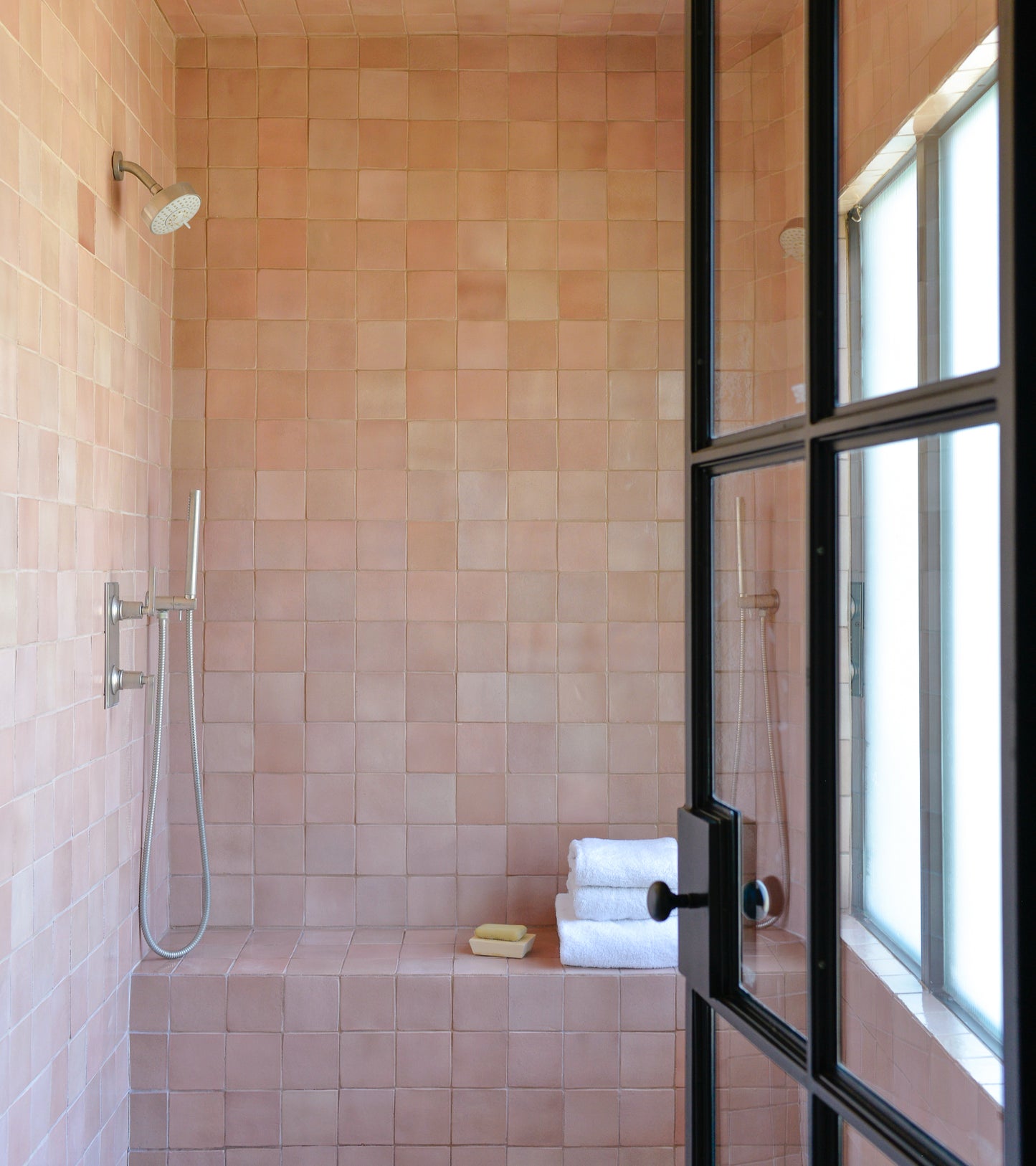 A modern shower features Zia Tile’s Posada 4x4 handmade pink Cotto tiles, a rainfall and handheld shower, built-in bench, and white towels with soap, all near a glass door and window.