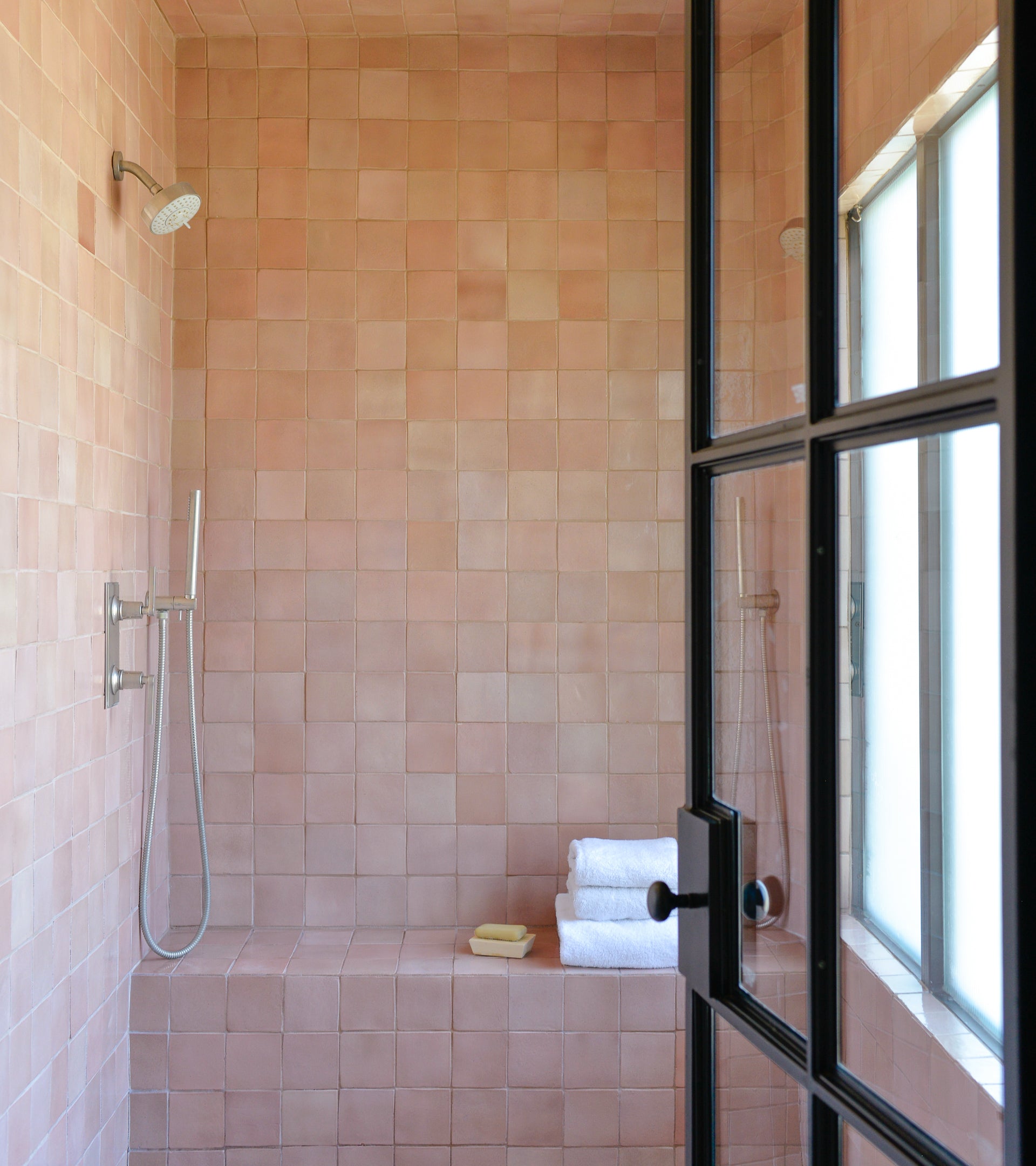 A modern shower features Zia Tile’s Posada 4x4 handmade pink Cotto tiles, a rainfall and handheld shower, built-in bench, and white towels with soap, all near a glass door and window.
