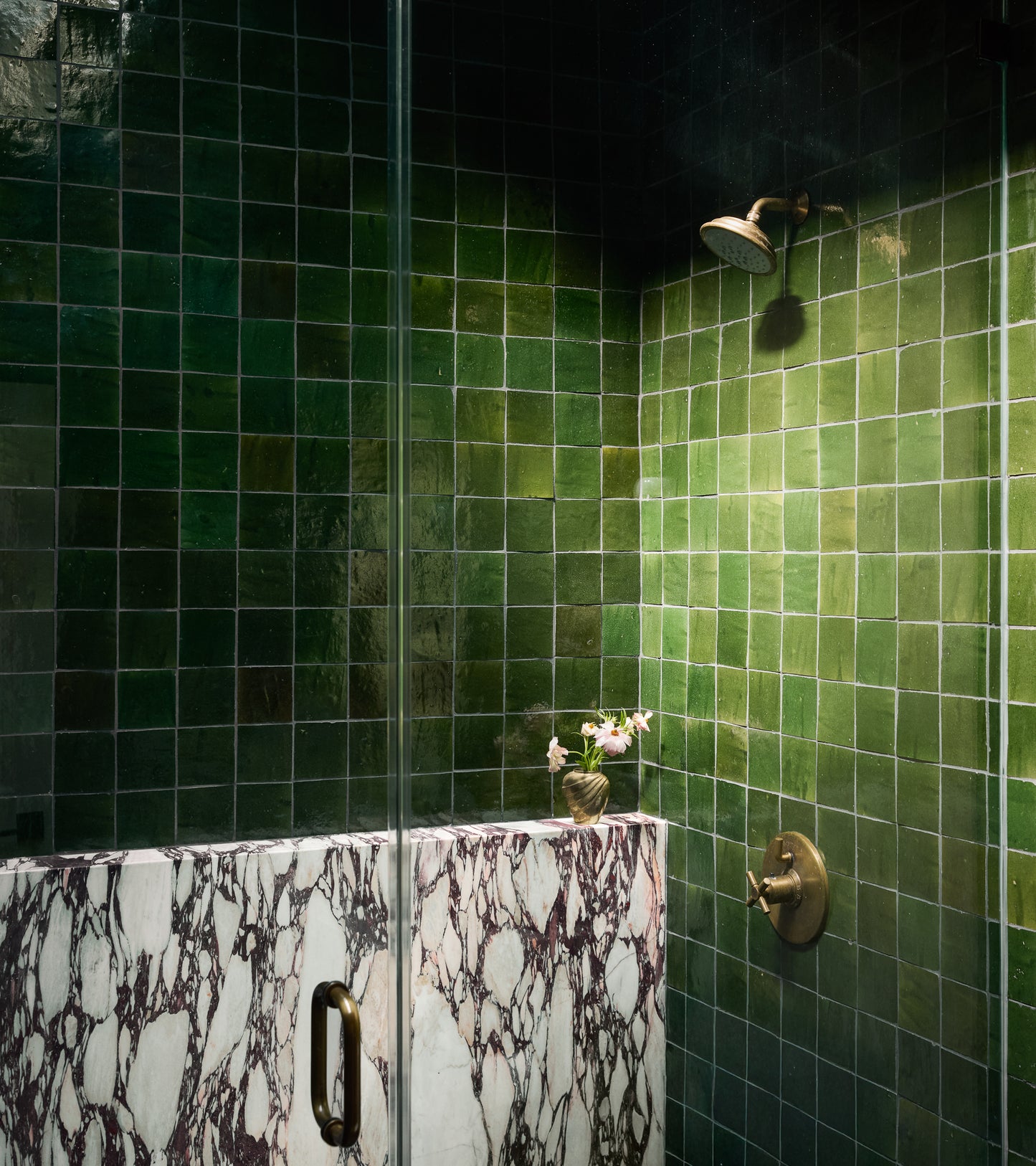 A shower featuring Racing Green 4x4 tiles, brass fixtures, and a white marble half-wall with dark veining; a small flower vase rests on the marble ledge.