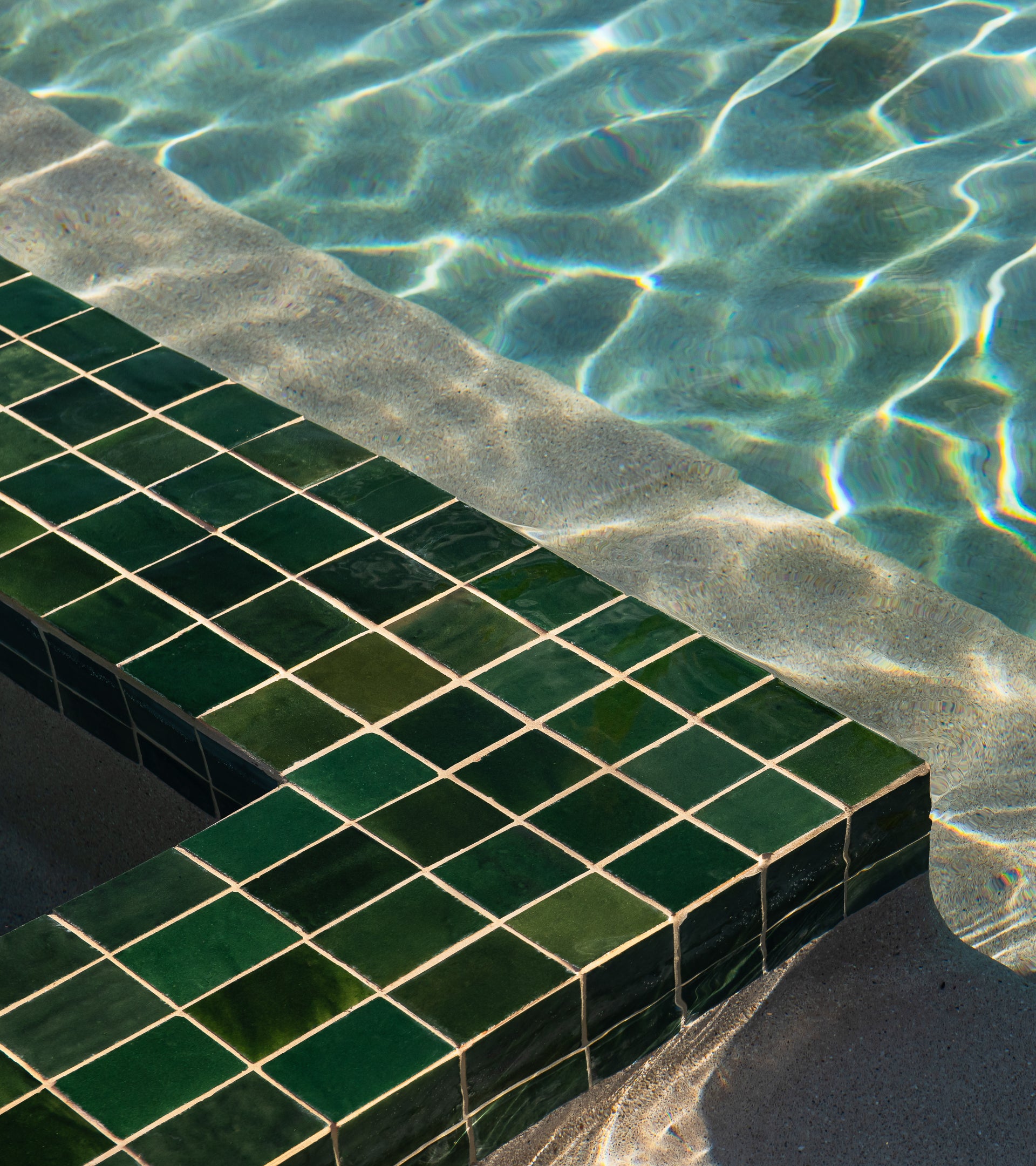 Sunlight reflects on the clear water of a swimming pool, casting patterns on vendor-unknown's Racing Green 4x4 tiles that line the green-tiled pool steps partially submerged in water.