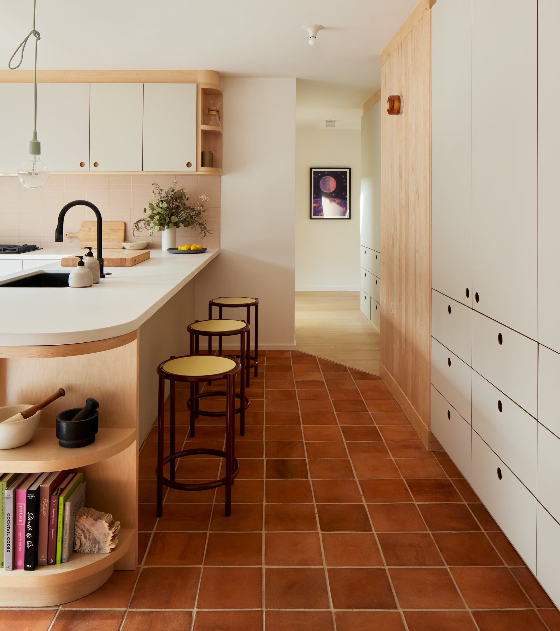 Modern kitchen with Zia Tile 8x8 Square + Red Clay terracotta floor tiles, light wood and white cabinets, open shelves with books, a vase of flowers on the counter, two stools, and a hallway with art evoking San Miguel de Allende style.