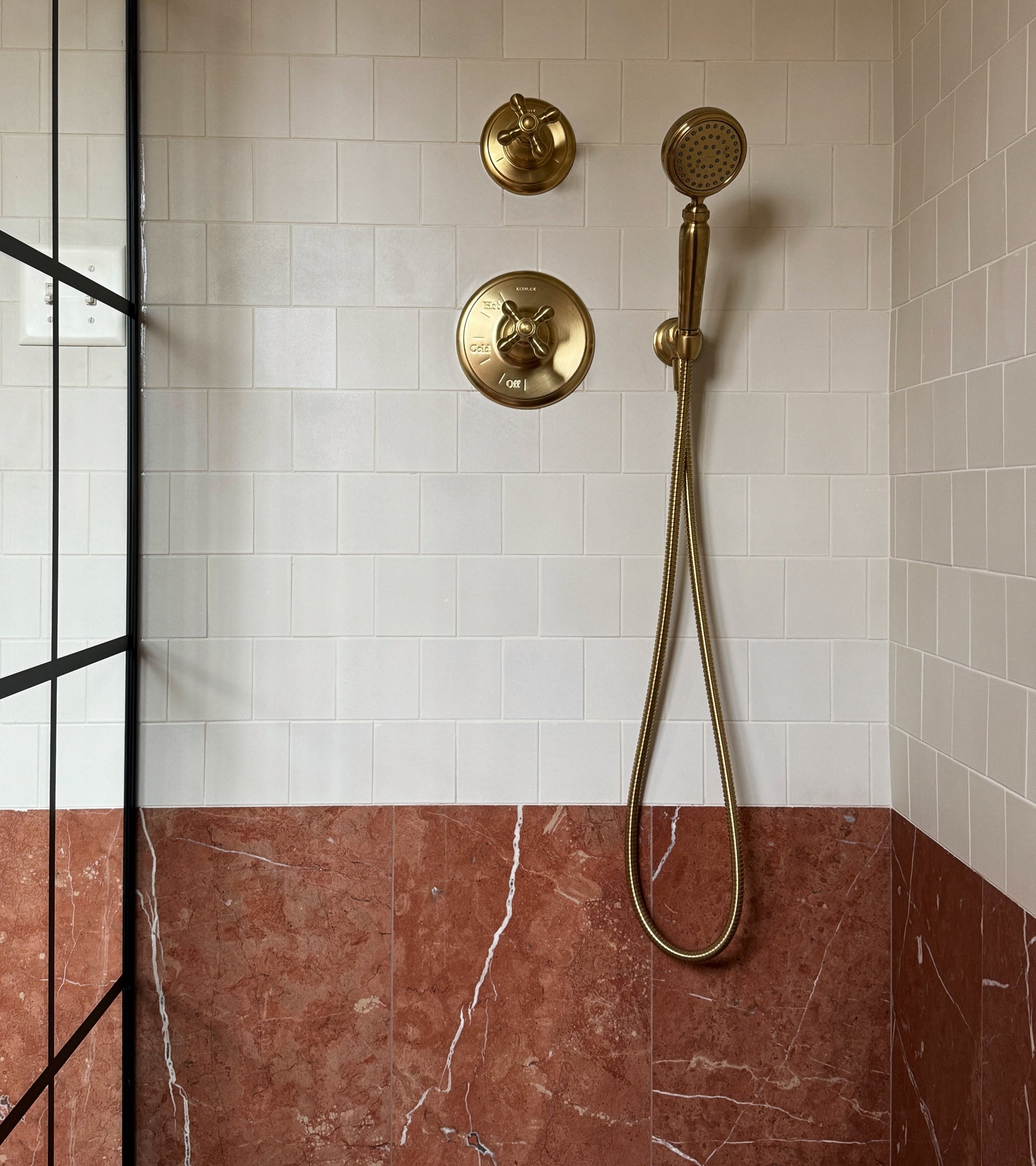 A modern shower features white handmade upper wall tiles and Zia Tile’s Rosso Alicante 12x24 reddish-brown marble lower tiles. Gold fixtures—handheld showerhead, two knobs—and a black-framed glass partition complete the space.