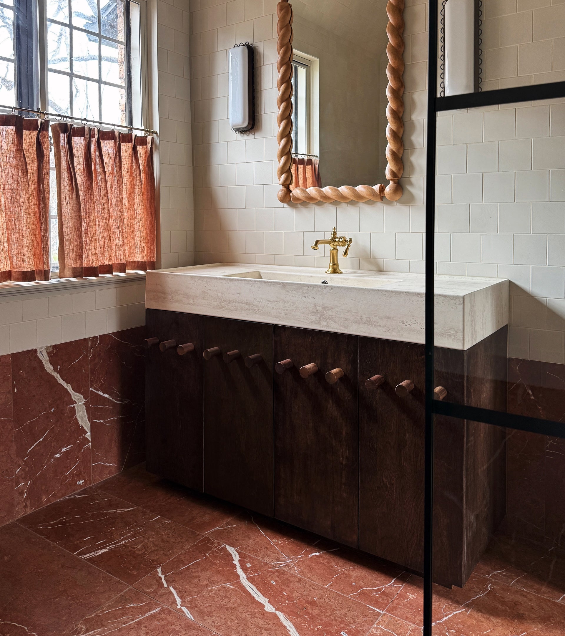 A bathroom with Rosso Alicante 12x24 red marble floor and lower walls by Zia Tile, a large rectangular sink with gold faucet, dark wood knob-handle cabinets, a twisted frame mirror, and a window with red curtains.
