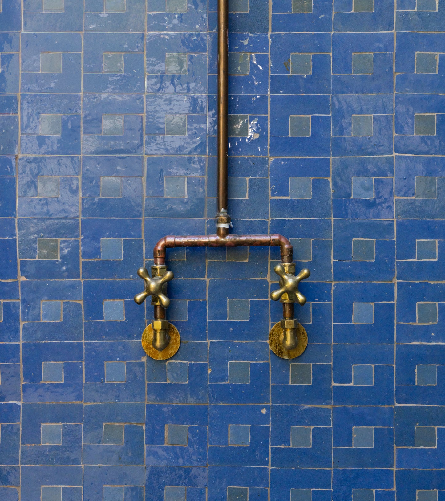 The Rubric 7 by Zia Tile features two brass faucet handles linked by copper pipes, mounted on a wall adorned with neatly arranged blue Moroccan tiles.