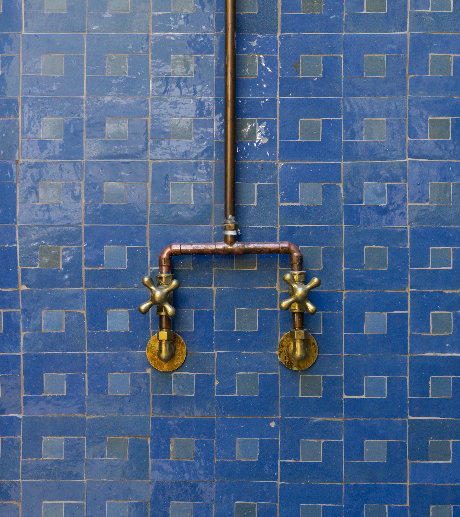 The Rubric 7 by Zia Tile features two brass faucet handles linked by copper pipes, mounted on a wall adorned with neatly arranged blue Moroccan tiles.