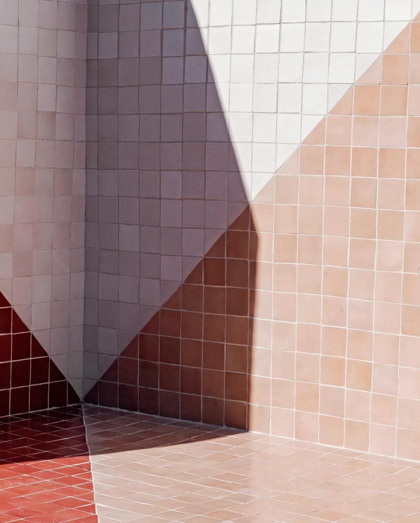 The wall and floor feature Zia Tiles Posada 4x4 tiles in terra cotta shades of red, pink, beige, and white. A sharp diagonal shadow adds contrast between light and dark areas across the geometric surface.