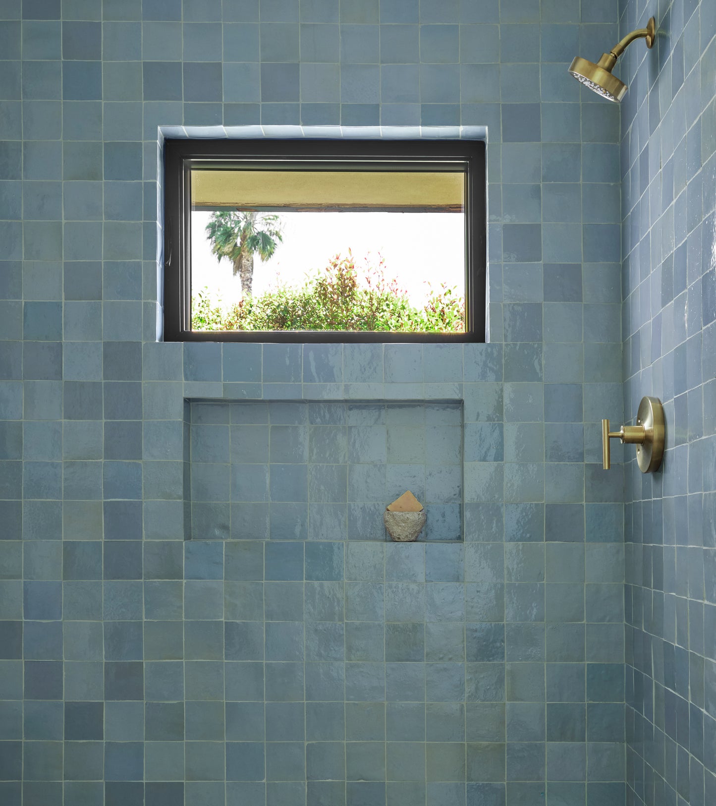 Shower with glossy Superior Blue 4x4 tiles by vendor-unknown, a built-in shelf for soap, a window with greenery and palm tree views, plus a gold showerhead and handle on the right wall.