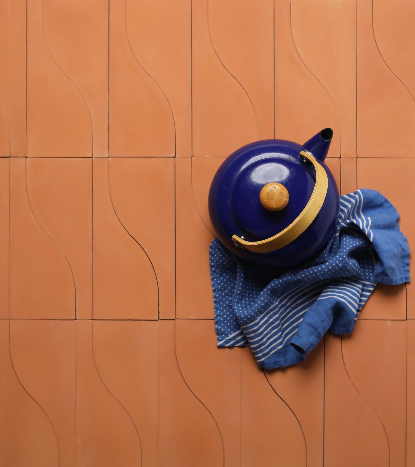 A blue teapot with a wooden handle sits on a folded blue and white cloth atop vendor-unknown’s Nouveau Terra Cotta tiles, featuring a wavy grid pattern—ideal inspiration for your next kitchen backsplash.