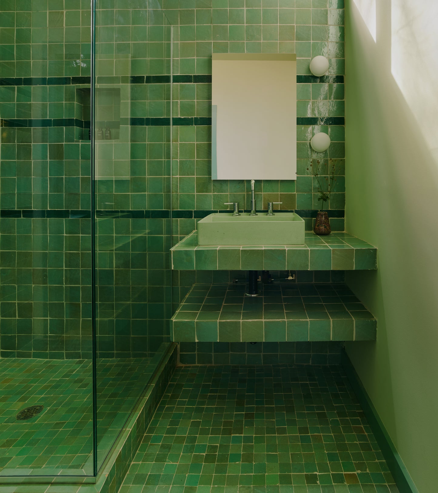 A modern bathroom features Tidepool 4x4 tiles in green on the floor, walls, and sink, complemented by a glass shower, rectangular mirror, two round wall lights, and a small vase with greenery on the floating counter.