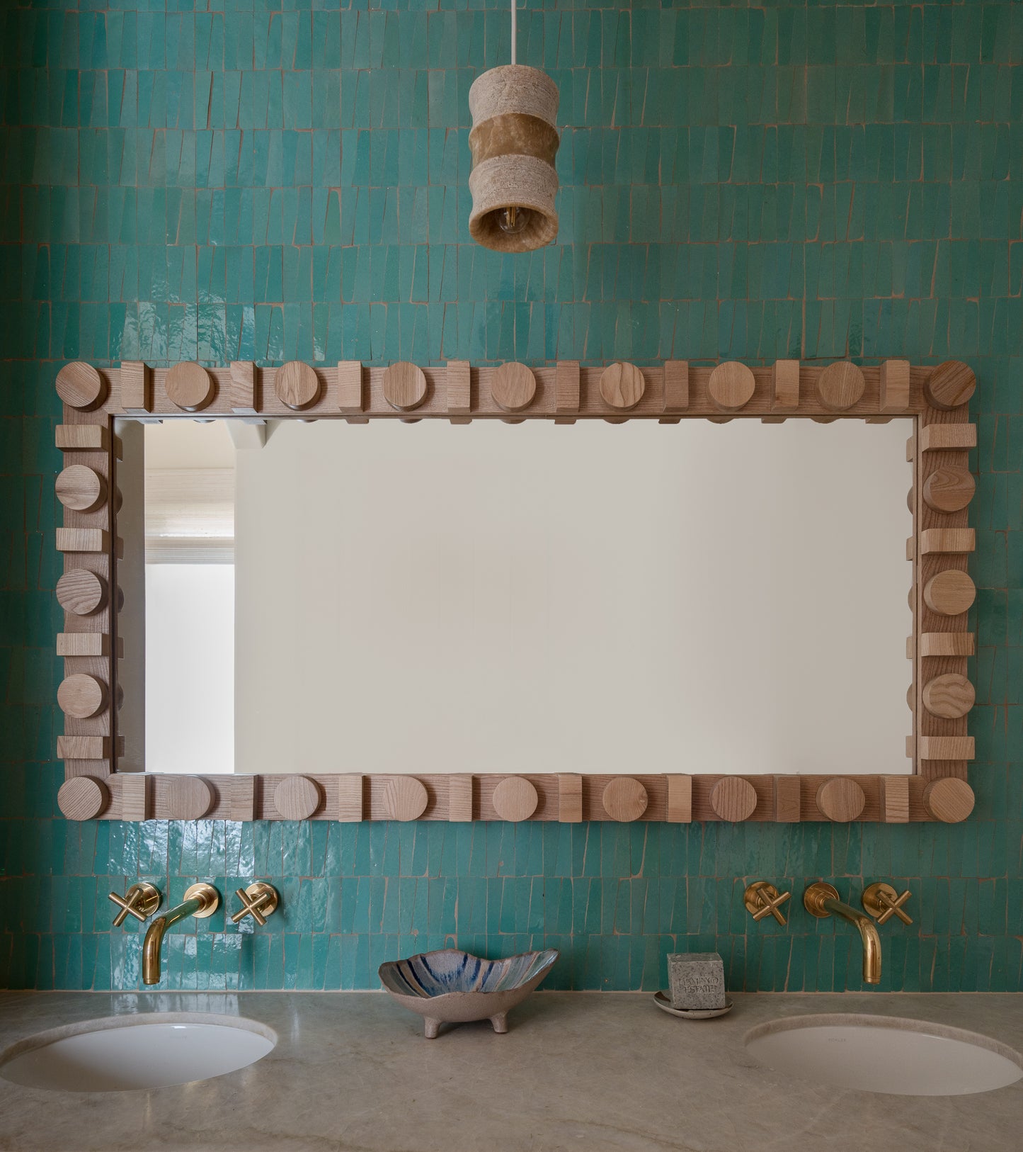 A bathroom featuring Tulum Trapezoid tiles by Zia Tile in turquoise, a large geometric wood-framed mirror, twin sinks with gold faucets, a shell dish and soap bar, and a sculptural pendant light overhead.