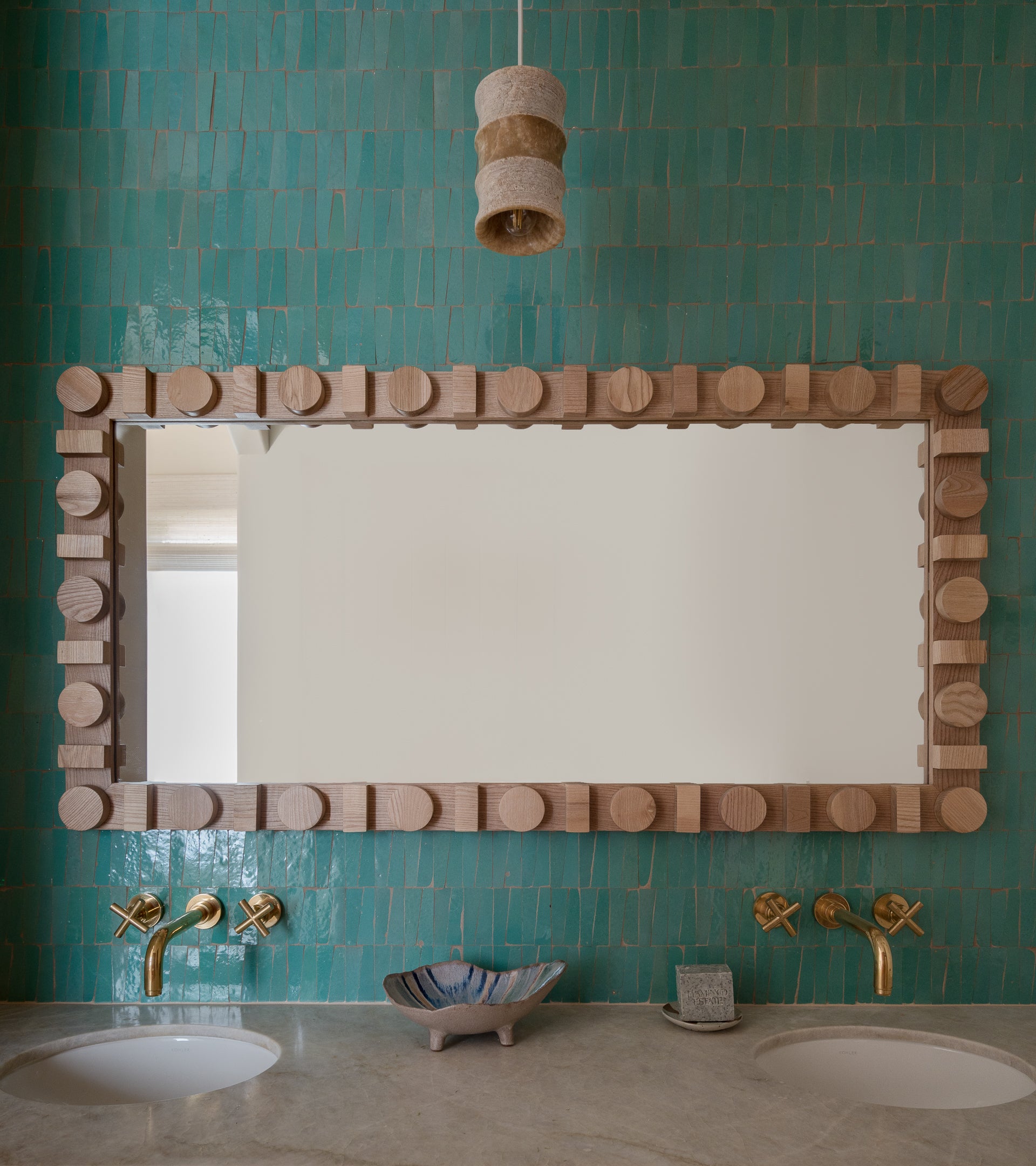 A bathroom featuring Tulum Trapezoid tiles by Zia Tile in turquoise, a large geometric wood-framed mirror, twin sinks with gold faucets, a shell dish and soap bar, and a sculptural pendant light overhead.