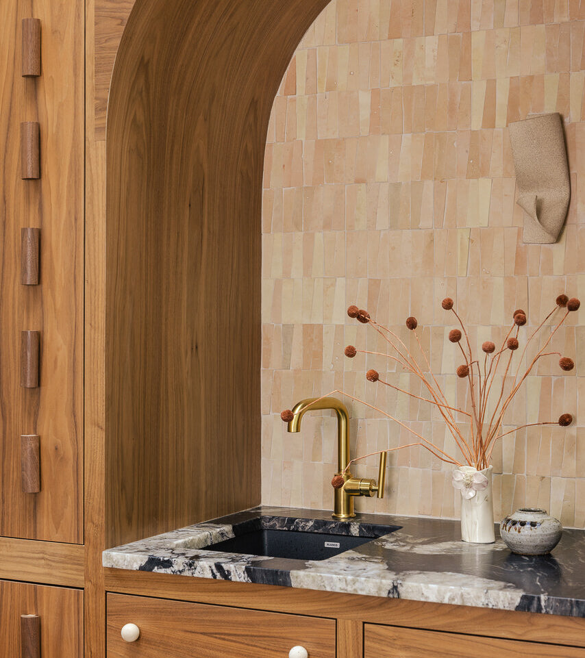 A small kitchen sink area features a gold faucet set in black marble, surrounded by wood cabinetry. The backsplash showcases Unglazed Natural Trapezoid tiles. By the sink sits a white vase holding brown branches.
