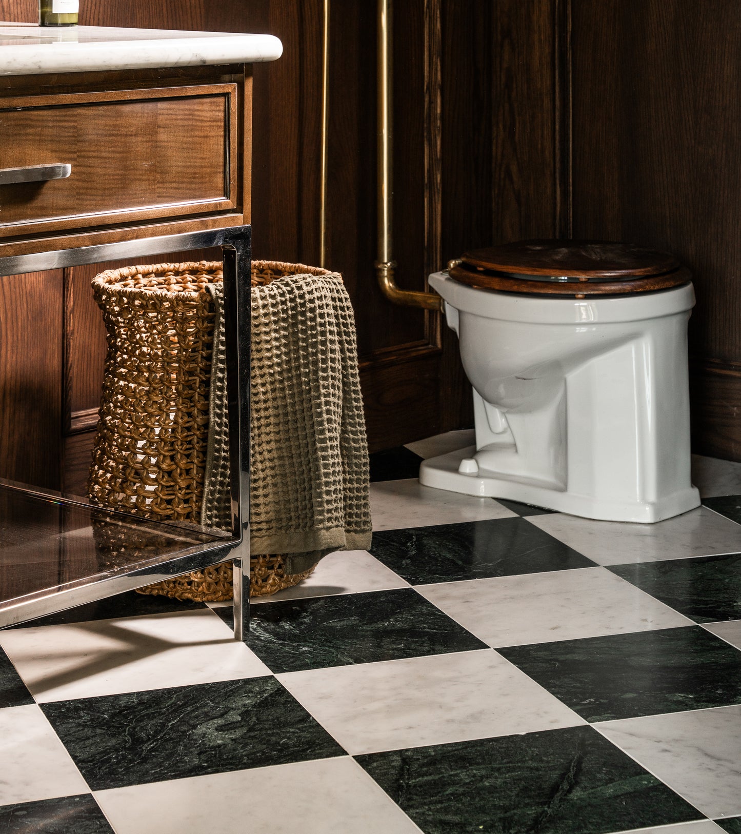 A bathroom featuring Zia Tile's Verde Alpi 12x12 tiles, a white toilet with a dark wooden seat, a wicker basket with a textured towel, and a metal-framed vanity next to a wood-paneled wall.