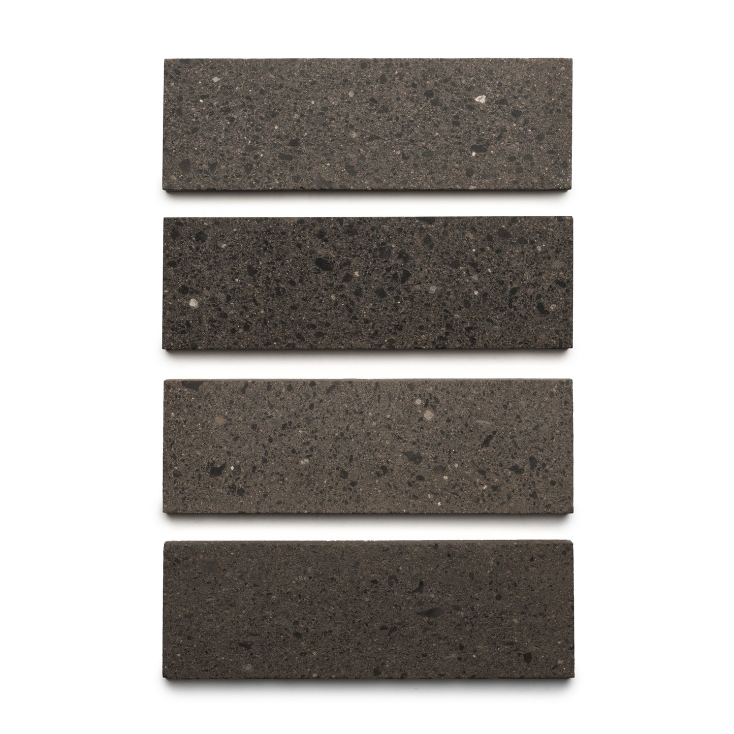 Four Volcan 4x12 rectangular tiles by Zia Tile, in dark gray terrazzo with earth-toned aggregates, are arranged vertically in a straight column on a white background.