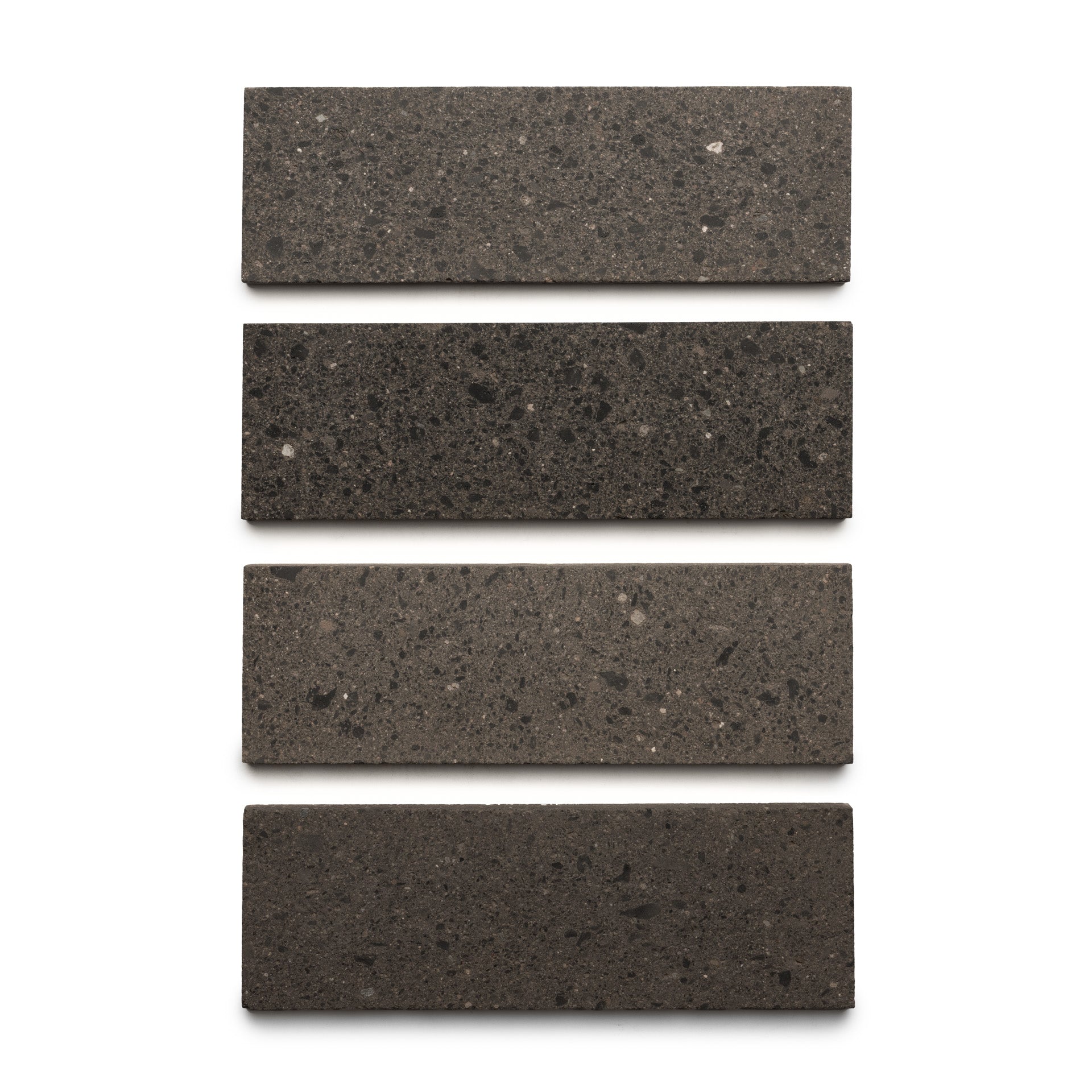 Four Volcan 4x12 rectangular tiles by Zia Tile, in dark gray terrazzo with earth-toned aggregates, are arranged vertically in a straight column on a white background.