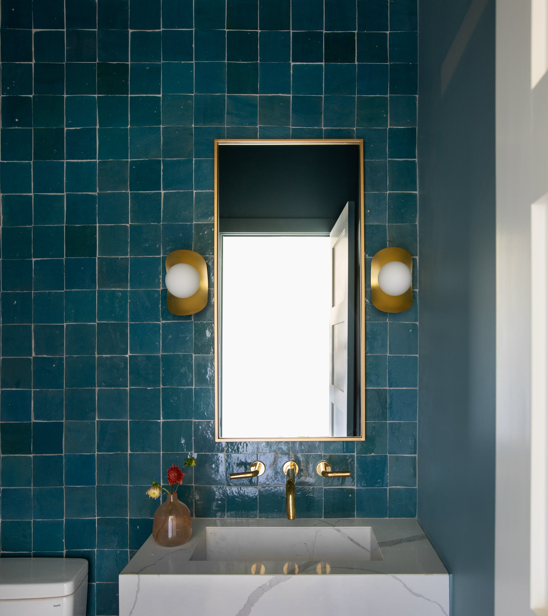 A modern bathroom features glossy teal Aegean 4x4 tiles by vendor-unknown, a rectangular mirror, two gold-based globe sconces, a white sink with gold fixtures, and a small vase with flowers on the counter.