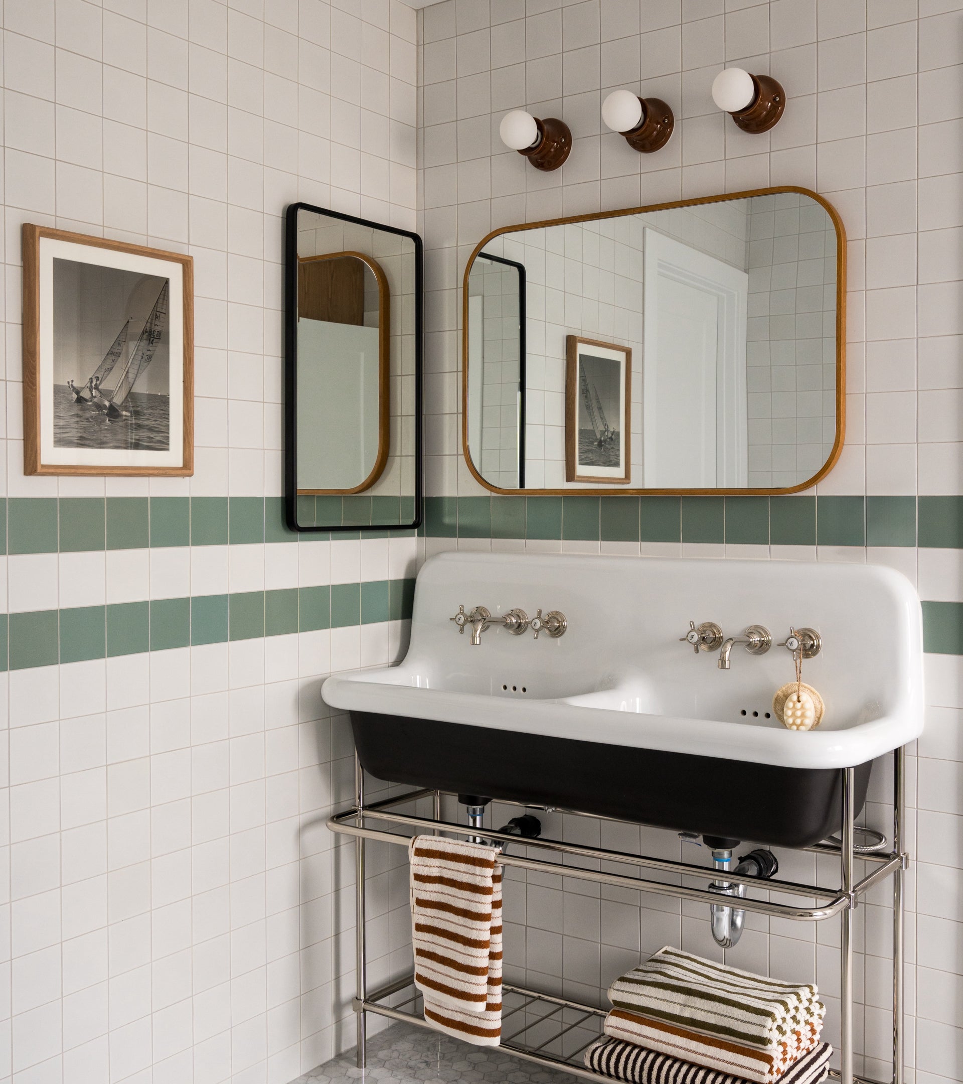 A vintage-style bathroom featuring Zia Tile's Alpha White 4x4 ceramics, a double white sink with chrome fixtures, two mirrors, striped towels, wall-mounted lights, and framed nautical art.
