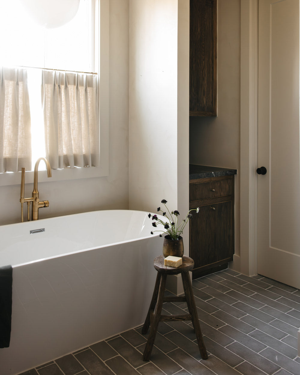 A modern bathroom features a white bathtub, gold faucet, and a small window with beige curtains. A wooden stool with a soap bar and flowers sits beside the tub on Zia Tile’s Basilica 4x12 + Honed limestone flooring.