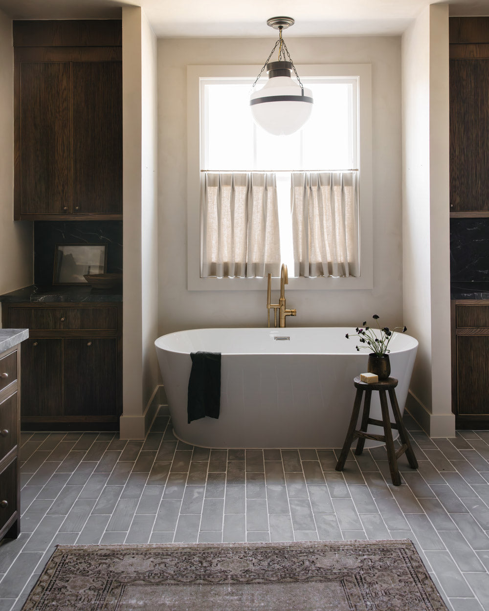 A modern bathroom features a freestanding white bathtub below a window with beige curtains, dark wood cabinets, and Zia Tile's Basilica 4x12 Honed tiles. A gold faucet, dark towel, rug, and floral stool add the finishing touches.