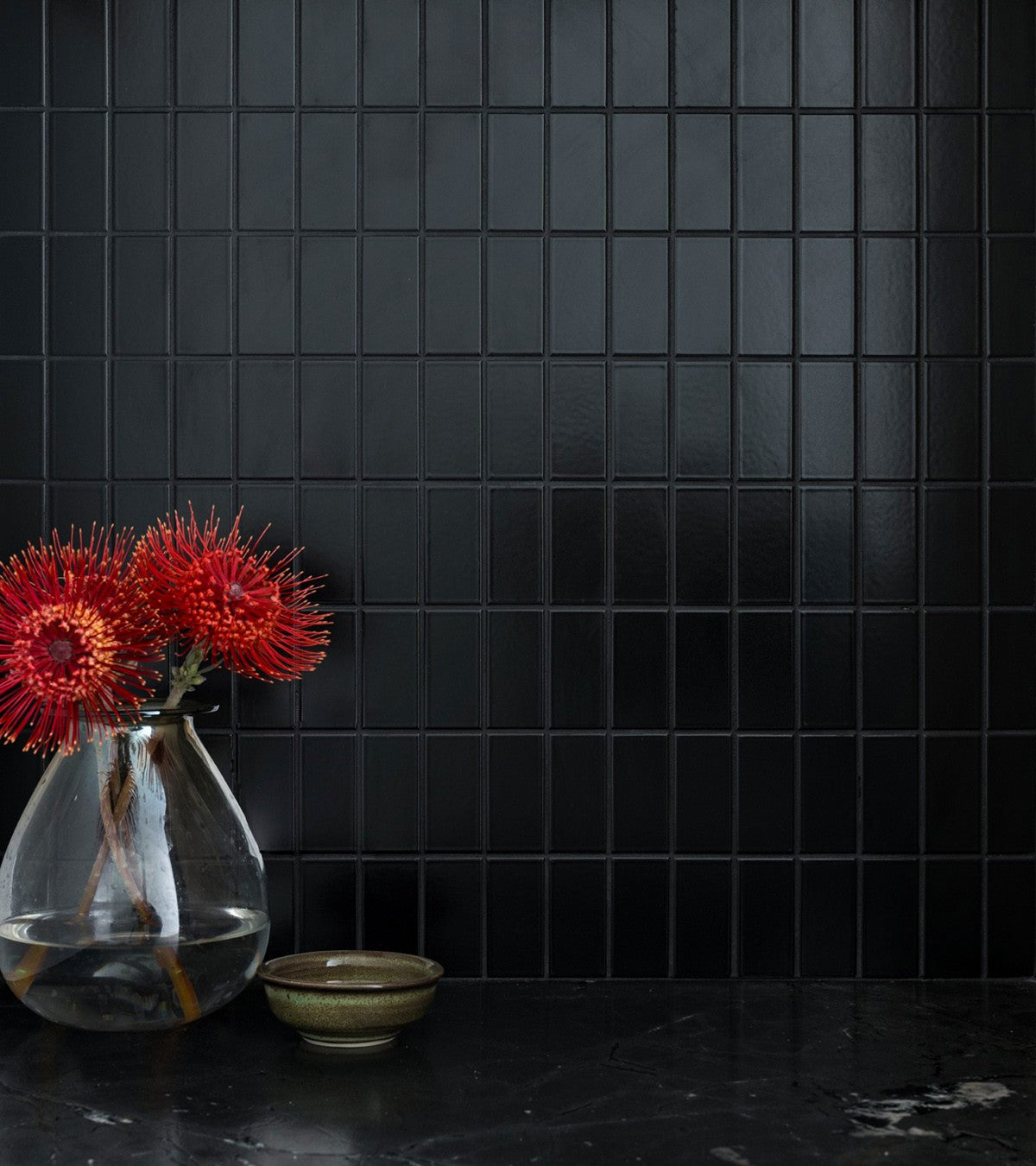 A glass vase with red-orange spiky flowers and water sits on a dark marble surface beside a small ceramic bowl, set against a wall of Zia Tile’s Cosmic Black 2x4 architectural grade ceramic tiles.