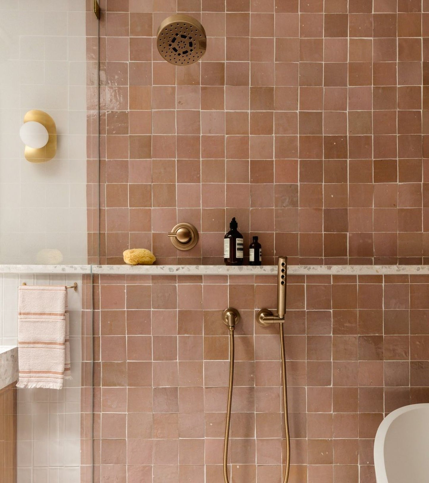 A modern shower featuring vendor-unknown's Desert Bloom 4x4 square pink tiles, brass fixtures, a marble shelf with bottles and a sponge, a striped towel on the left, and a round wall light reflected in the glass divider.