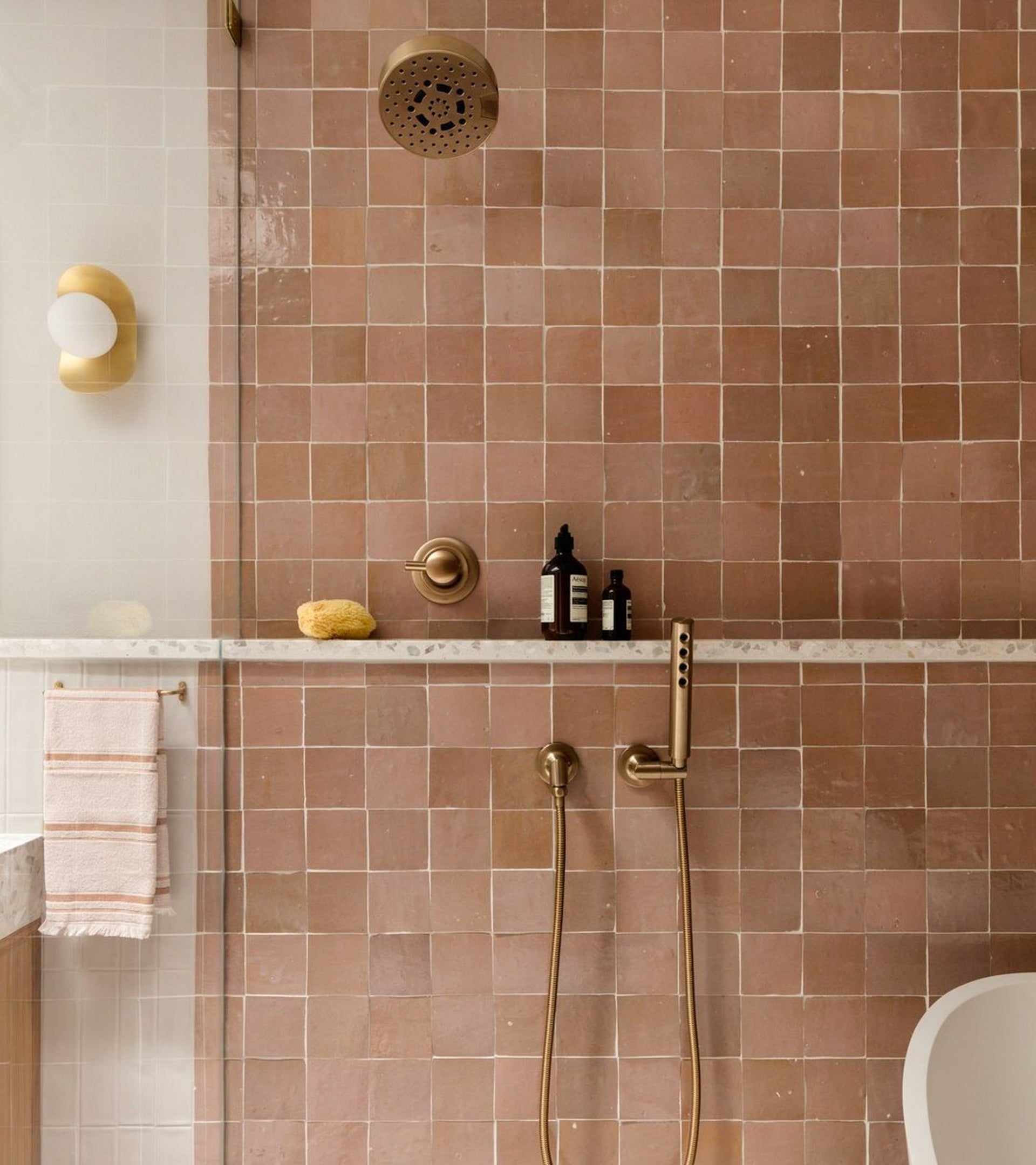 A modern shower featuring vendor-unknown's Desert Bloom 4x4 square pink tiles, brass fixtures, a marble shelf with bottles and a sponge, a striped towel on the left, and a round wall light reflected in the glass divider.
