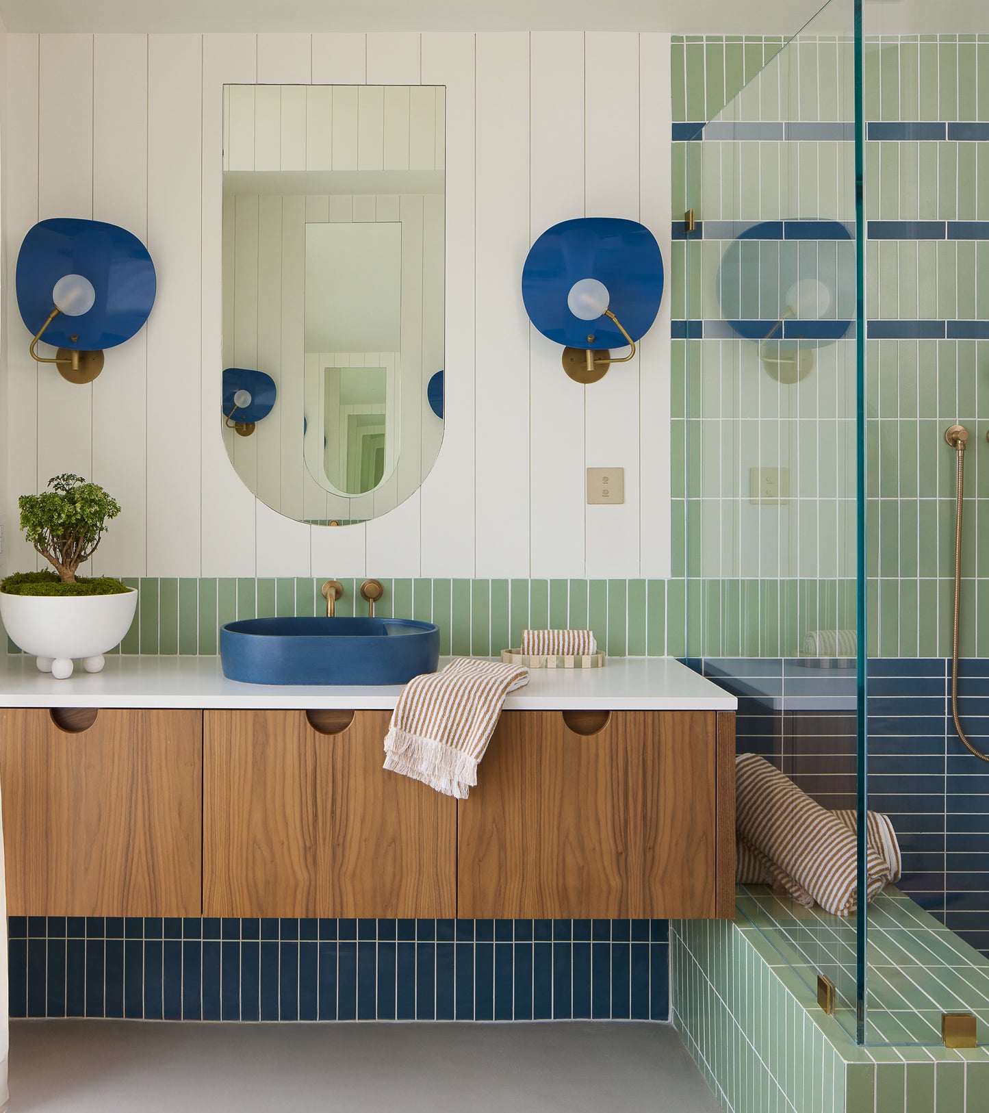 A modern bathroom features Zia Tile’s Eucalyptus 2x8 ceramics on the walls, a floating wood vanity, oval mirror, blue sink and sconces, a white planter with greenery, towels folded on the counter and in the walk-in glass shower.