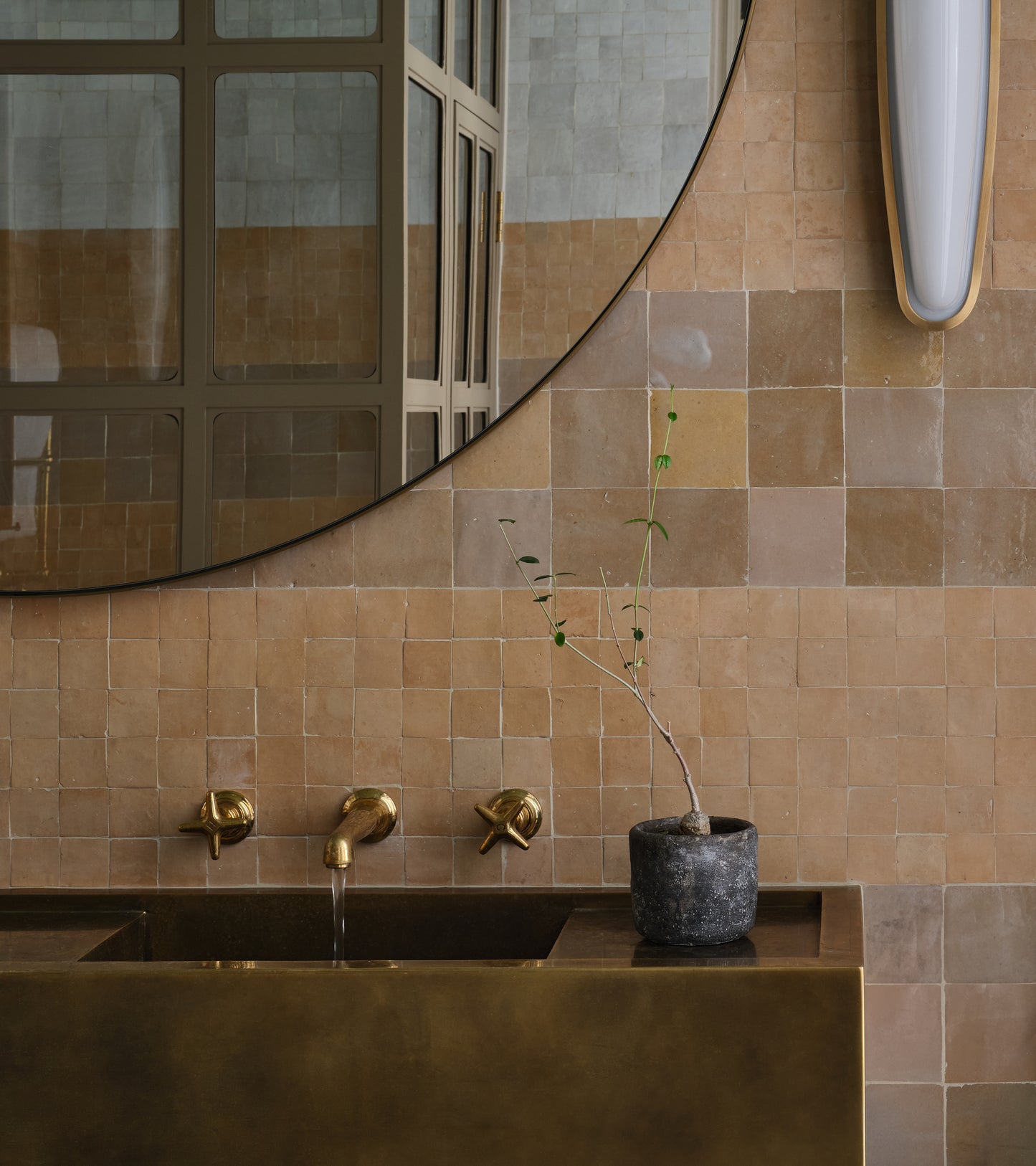A modern bathroom features Glazed Earth 4x4 tiles by vendor-unknown, a large round mirror, brass sink and fixtures, a small potted plant on the counter, and a white vertical light fixture mounted on the wall.