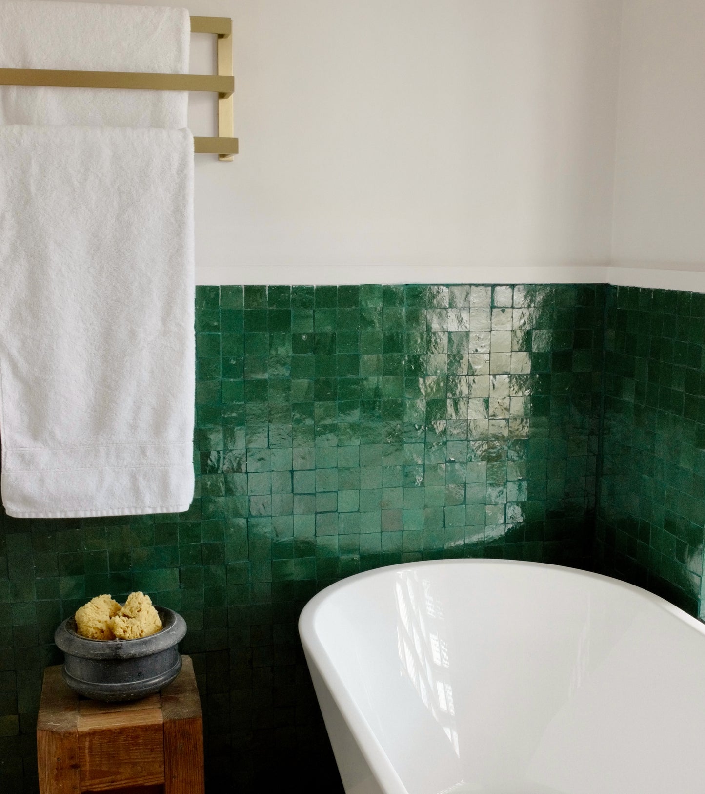 A white towel hangs on a gold rack above Jade 2x2 zellige tiles by Zia Tile. Below is a white bathtub, with a wooden stool nearby holding a bowl of sponges. The space feels clean and minimalistic.