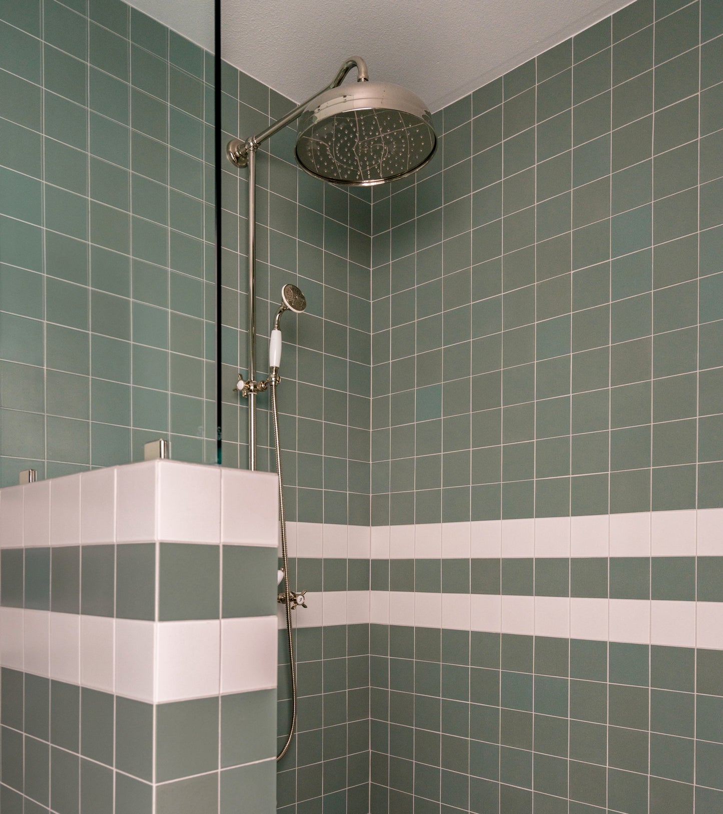 A modern shower features Zia Tile's Moonlight 4x4 green ceramics with two white horizontal stripes, a stainless steel rainfall showerhead with handheld attachment, and is partially enclosed by a tiled half-wall.