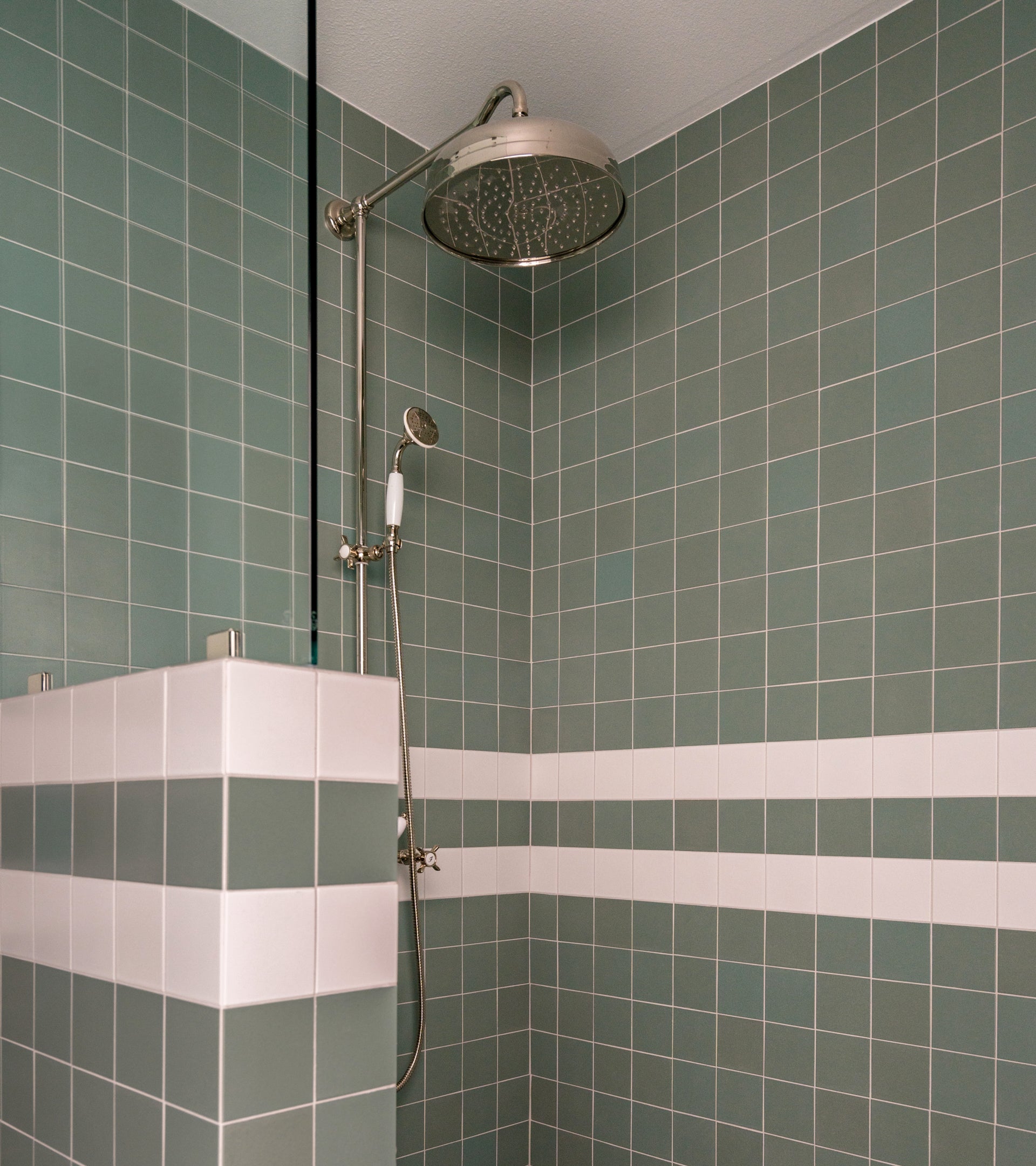 A modern shower features Zia Tile's Moonlight 4x4 green ceramics with two white horizontal stripes, a stainless steel rainfall showerhead with handheld attachment, and is partially enclosed by a tiled half-wall.