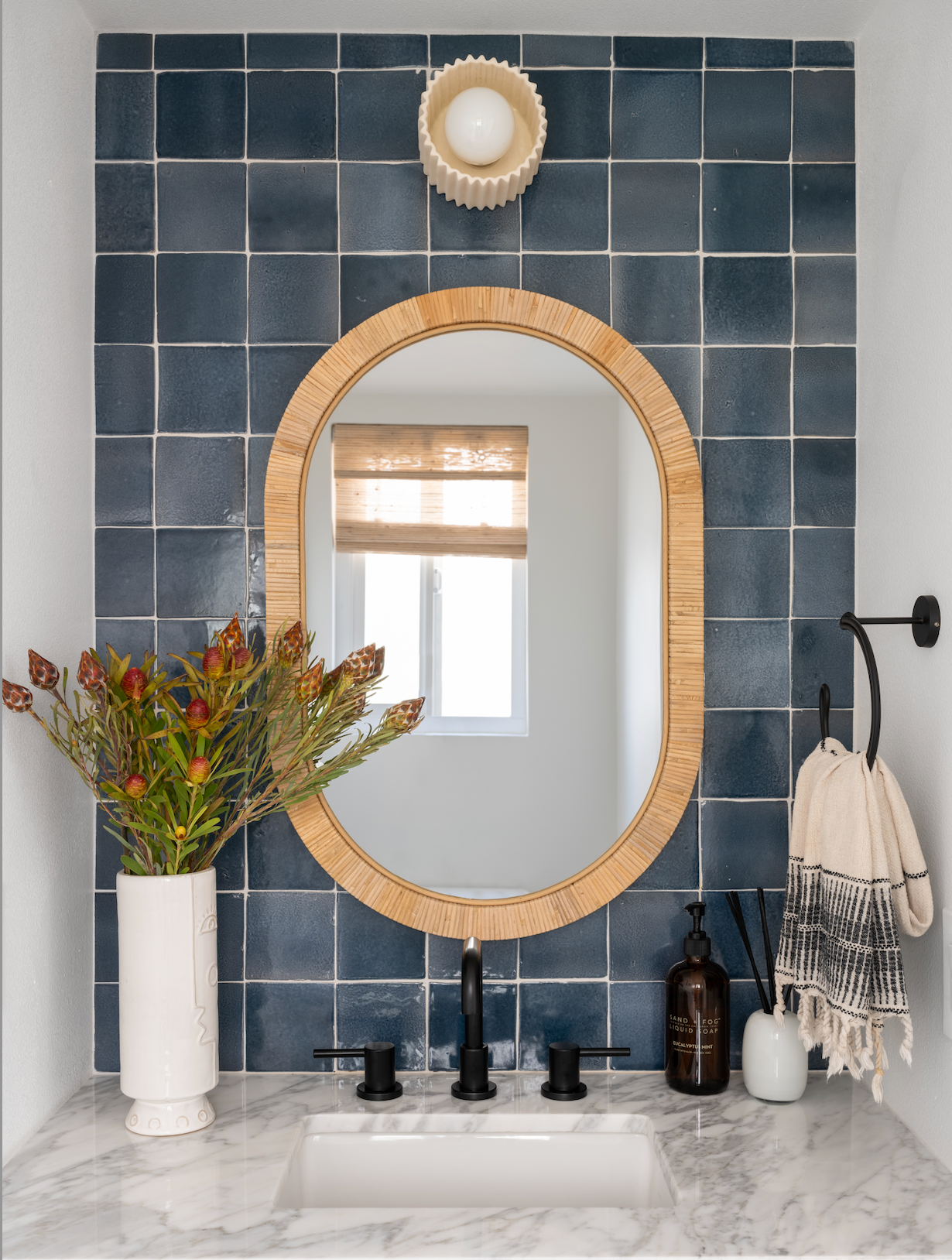 A bathroom vanity features a marble countertop, black faucet, oval woven-frame mirror, Zia Tile’s Piedra 4x4 Cotto ceramic blue square backsplash, vase of flowers, soap dispenser, and a hand towel next to the sink.